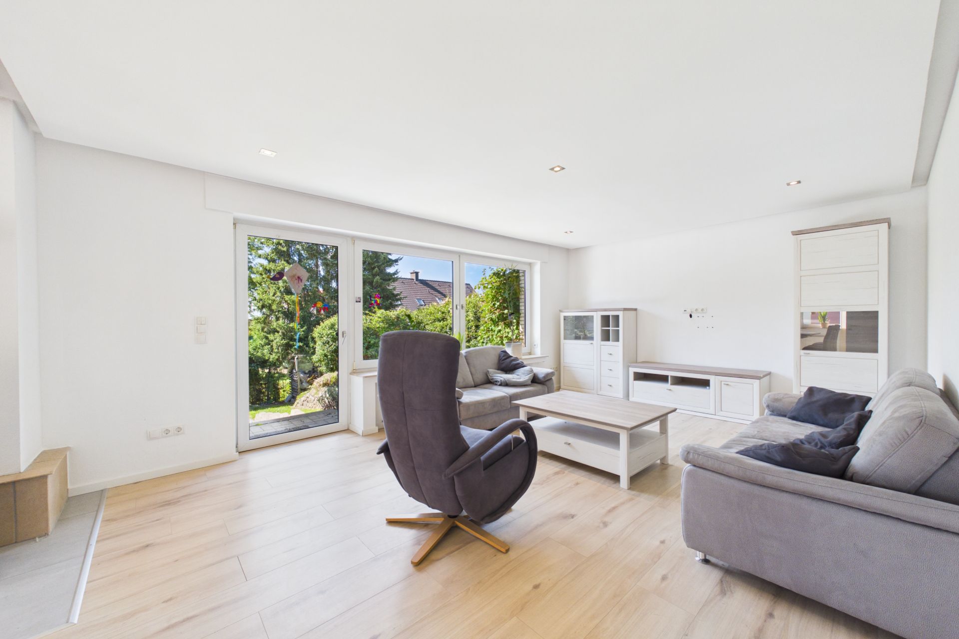Helles Wohnzimmer mit großen Fenstern, grauem Sofa, Sessel und Couchtisch auf Holzfußboden.