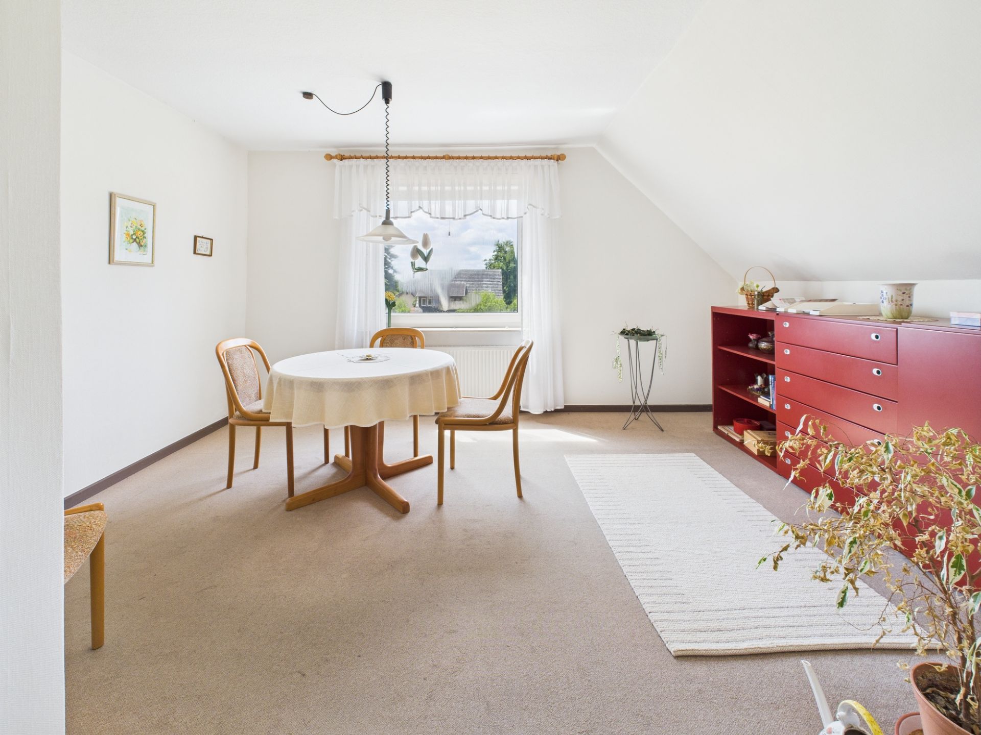 Helles Esszimmer mit rundem Tisch, drei Stühlen, roter Kommode und Fenster.