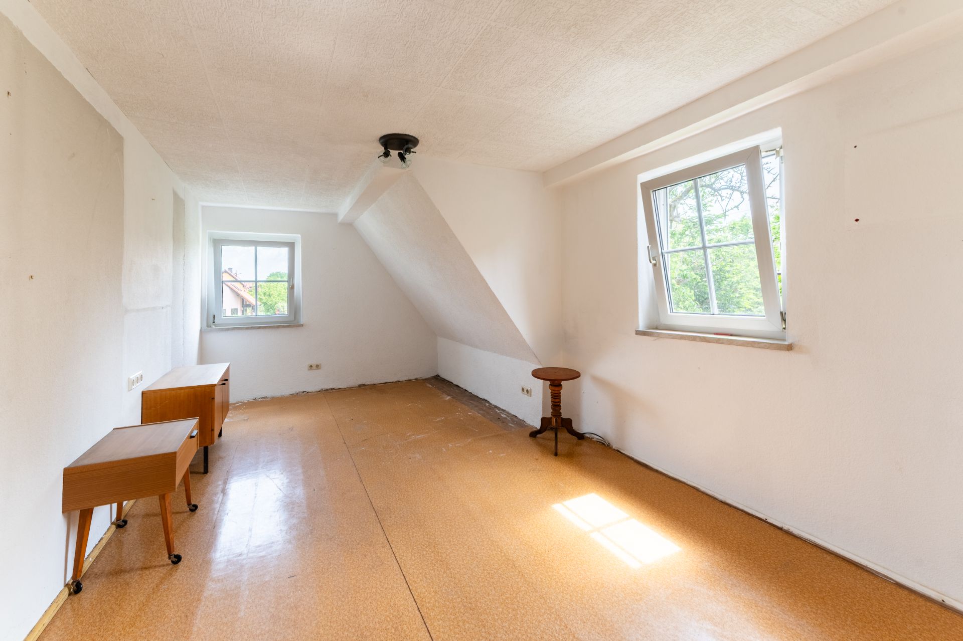 Leerer Raum mit zwei Fenstern, schräger Wand und wenigen kleinen Holzmöbeln.