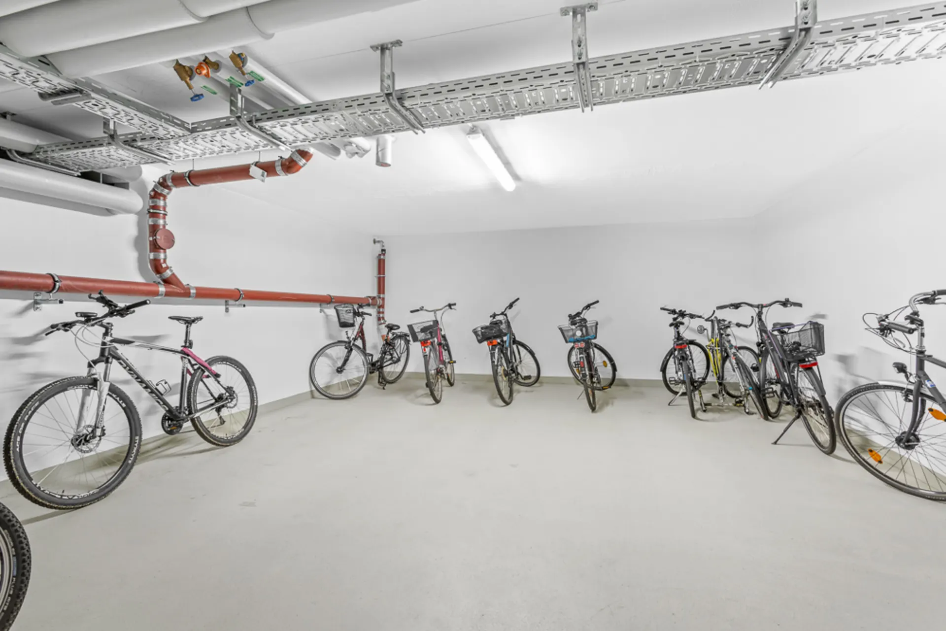Heller Fahrradkeller mit mehreren abgestellten Fahrrädern und sichtbaren Rohren an der Decke.