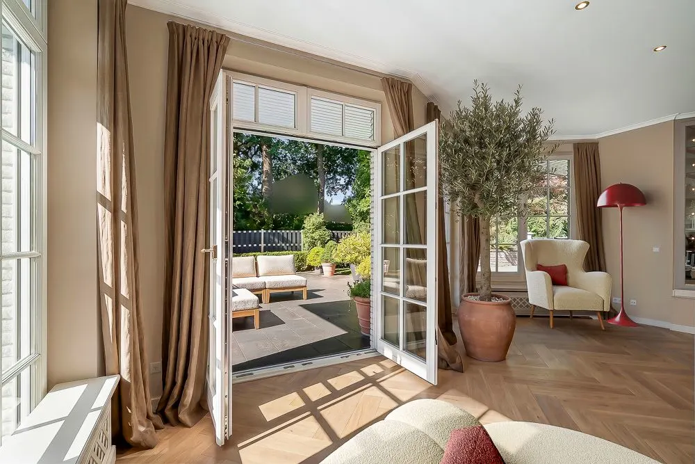 Wohnzimmer mit offener Terrassentür, Sessel, Stehlampe und Topfpflanze, Blick auf Terrasse mit Sitzbank.