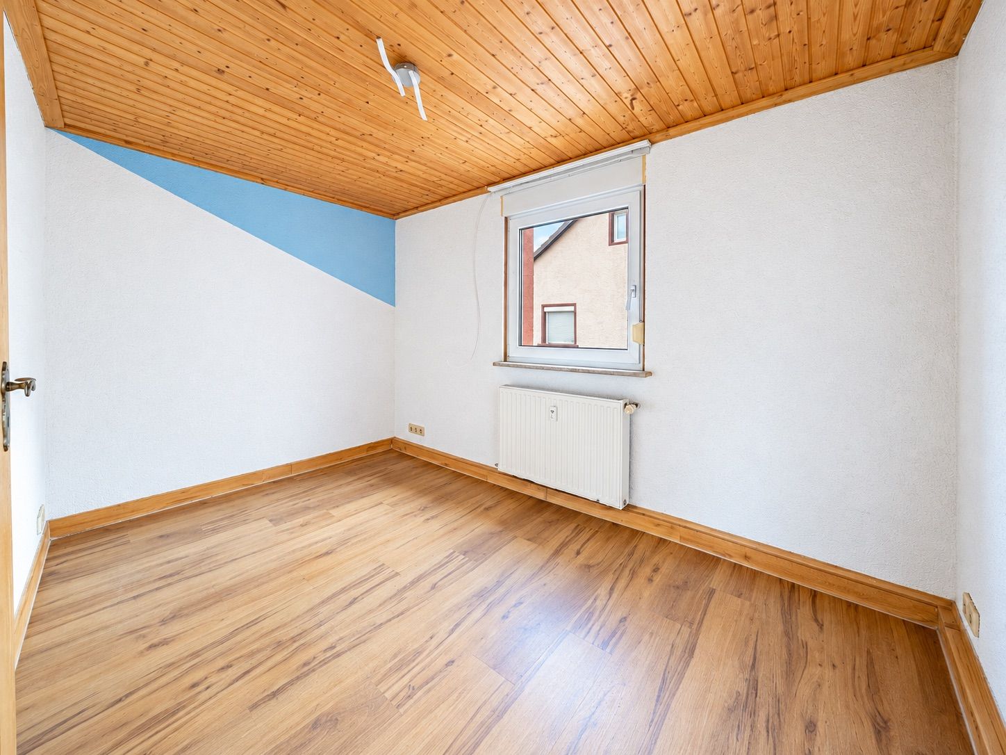 Leeres Zimmer mit Holzdecke, Laminatboden, Fenster und Heizkörper.