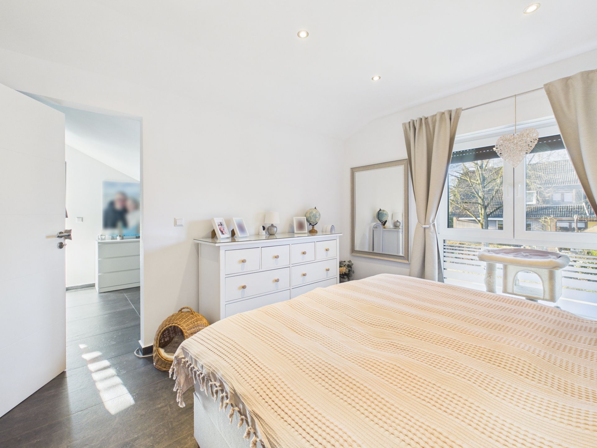 Helles Schlafzimmer mit großem Fenster, Vorhängen, Bett, Kommode und Spiegel.