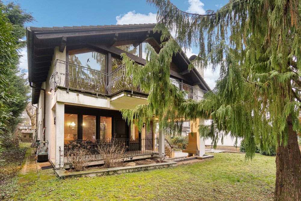 Zweistöckiges Wohnhaus mit Balkon, großen Fenstern und Garten, umgeben von Bäumen.
