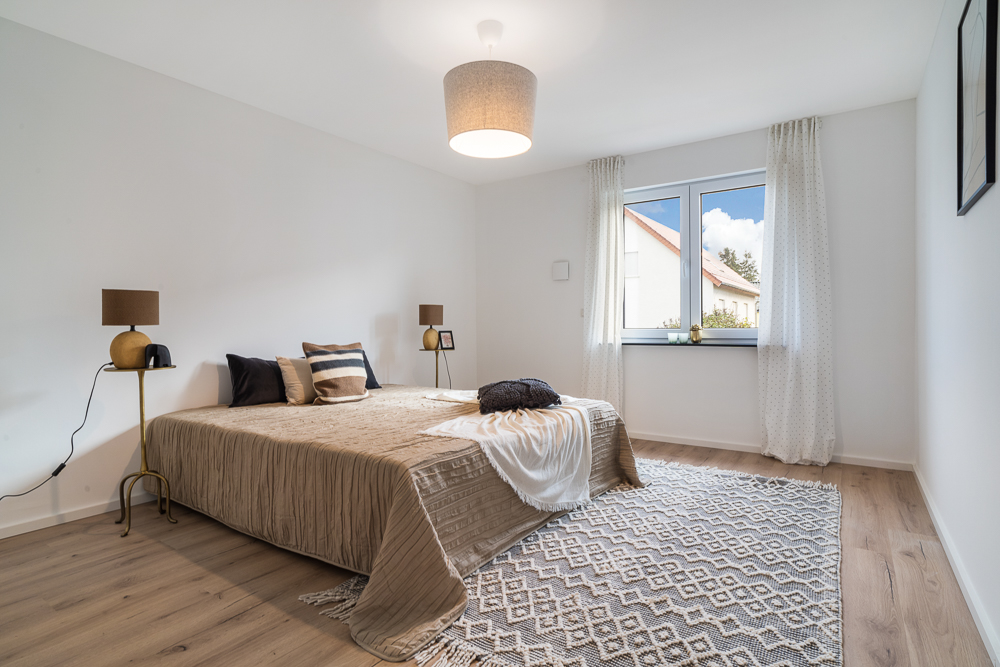 Helles Schlafzimmer mit Doppelbett, zwei Nachttischen und Fenster mit weißen Vorhängen.