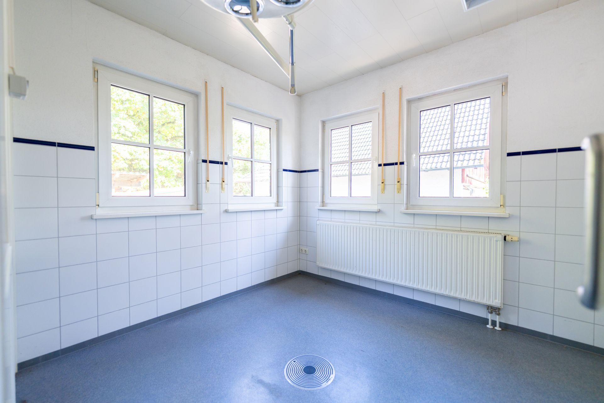 Heller Raum mit drei Fenstern, weißen Fliesen, Heizkörper und Abfluss im Boden.