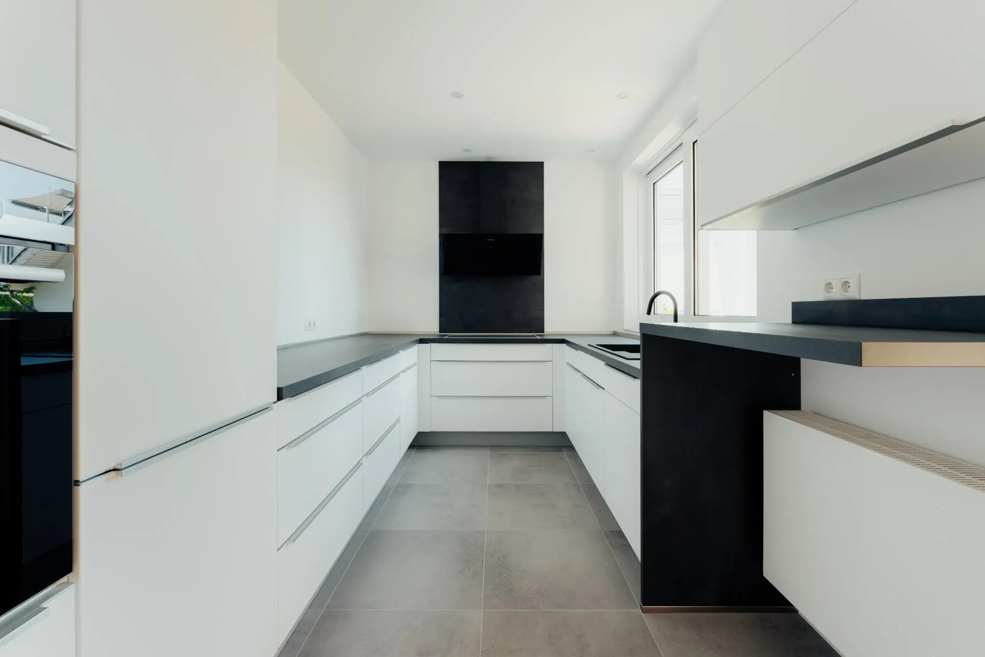 Moderne, weiße Einbauküche mit schwarzen Arbeitsflächen und großen Fenstern.