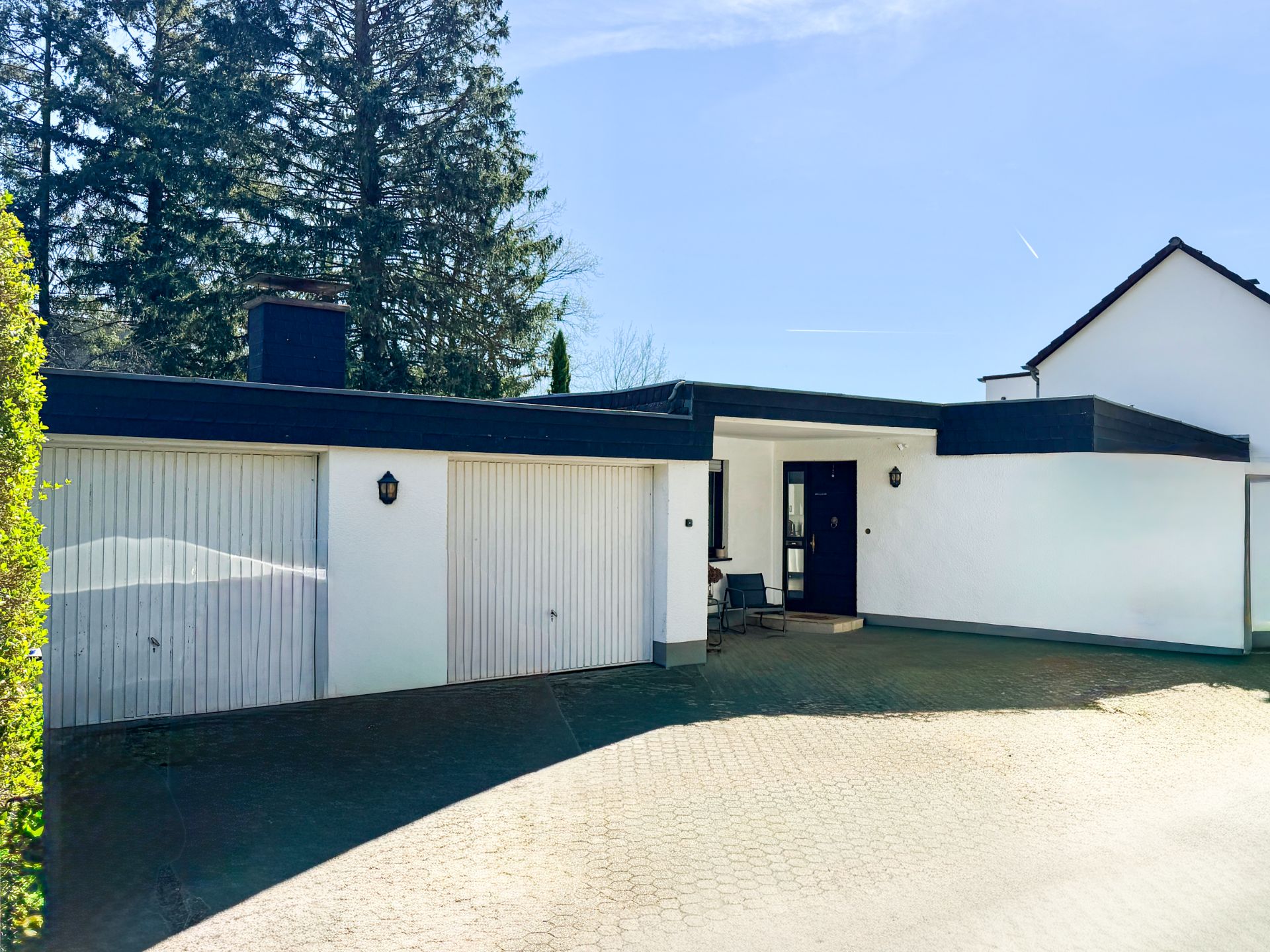 Einfamilienhaus mit Flachdach und zwei Garagentoren, gepflasterte Einfahrt, Bäume im Hintergrund.