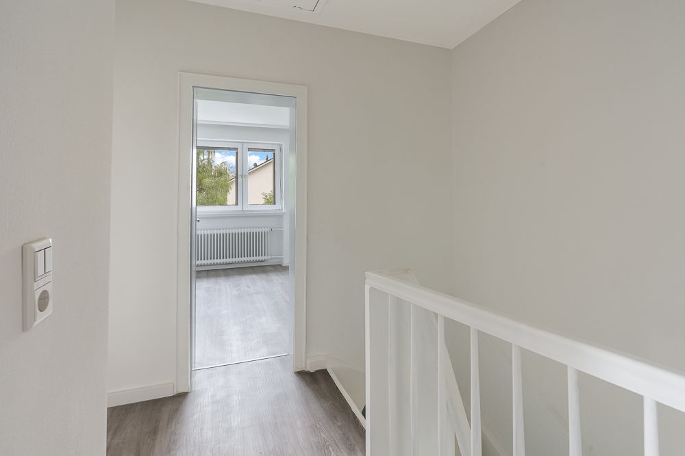 Heller Flur mit weißem Geländer und Blick in ein Zimmer mit großem Fenster.