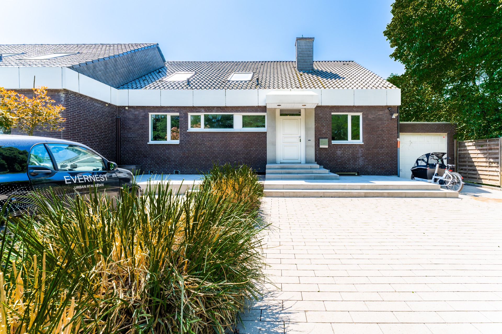 Moderne Backstein-Fassade mit weißer Eingangstür, vorne Pflastersteinweg und Auto links im Bild.