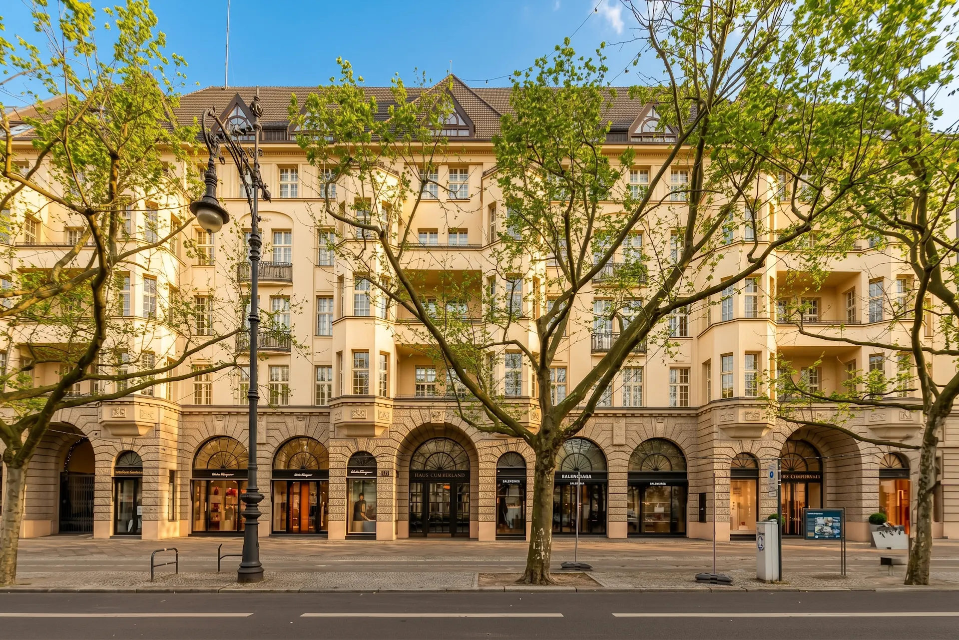 Mehrstöckiges Wohngebäude mit großen Fenstern und Arkaden, Bäume und Straße im Vordergrund.