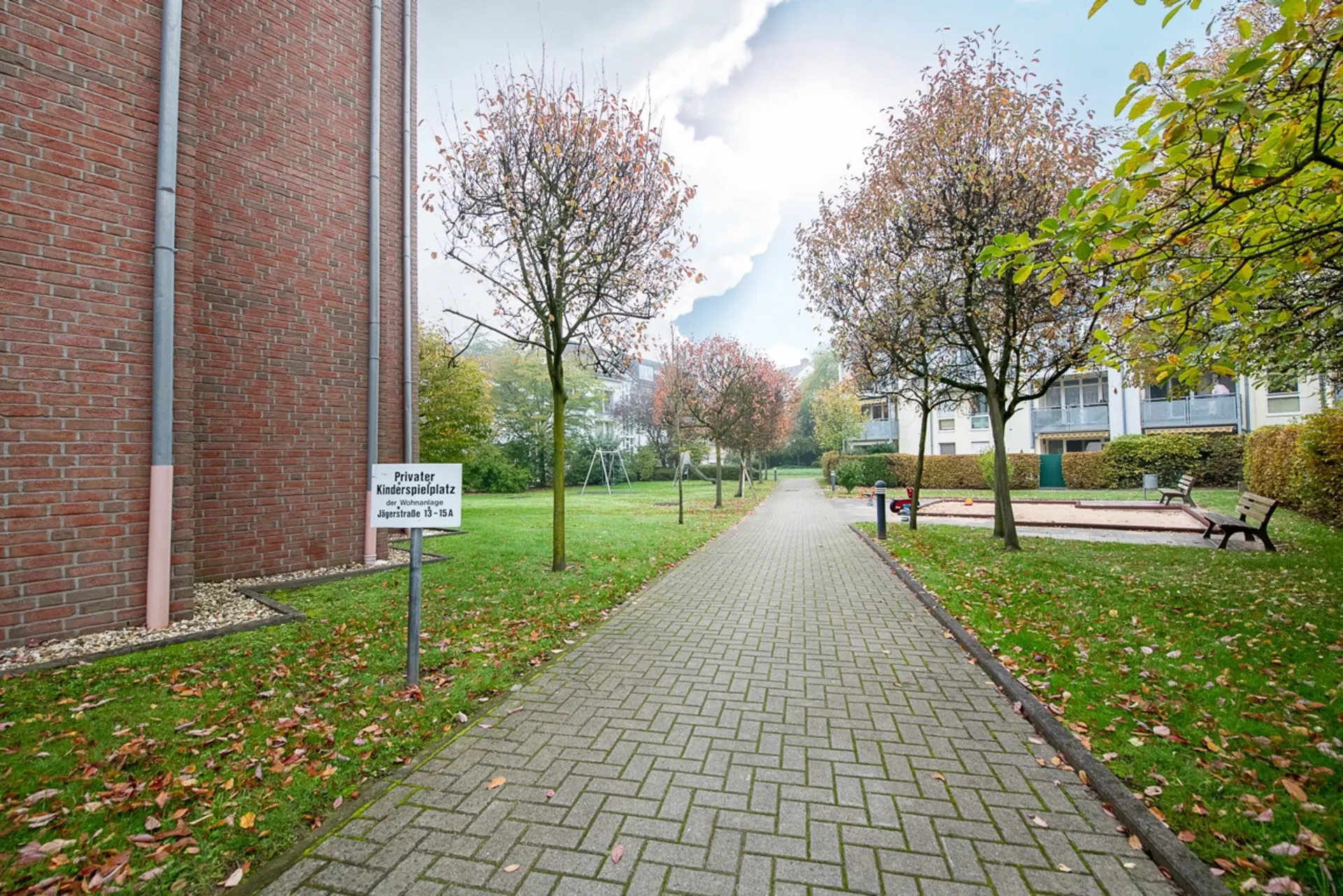 Gepflasterter Weg zwischen Bäumen mit Bänken und Sandkasten auf einer Wohnanlage.