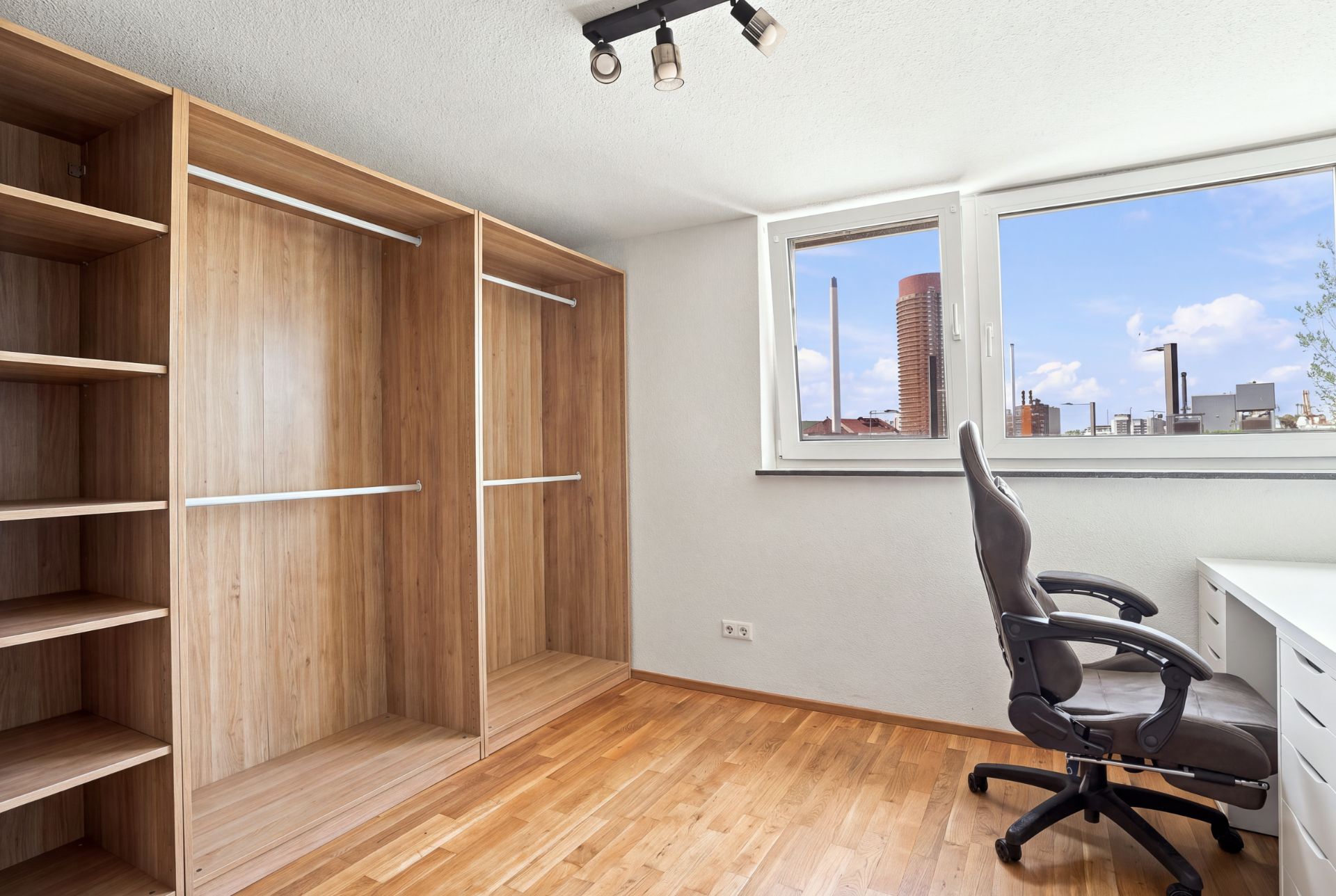 Helles Zimmer mit offenem Kleiderschrank, Schreibtisch, Bürostuhl und großem Fenster.