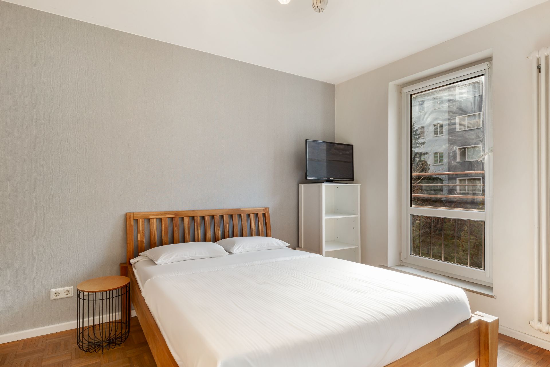 Schlafzimmer mit Holzbett, kleinem Tisch, Regal mit Fernseher und großem Fenster.