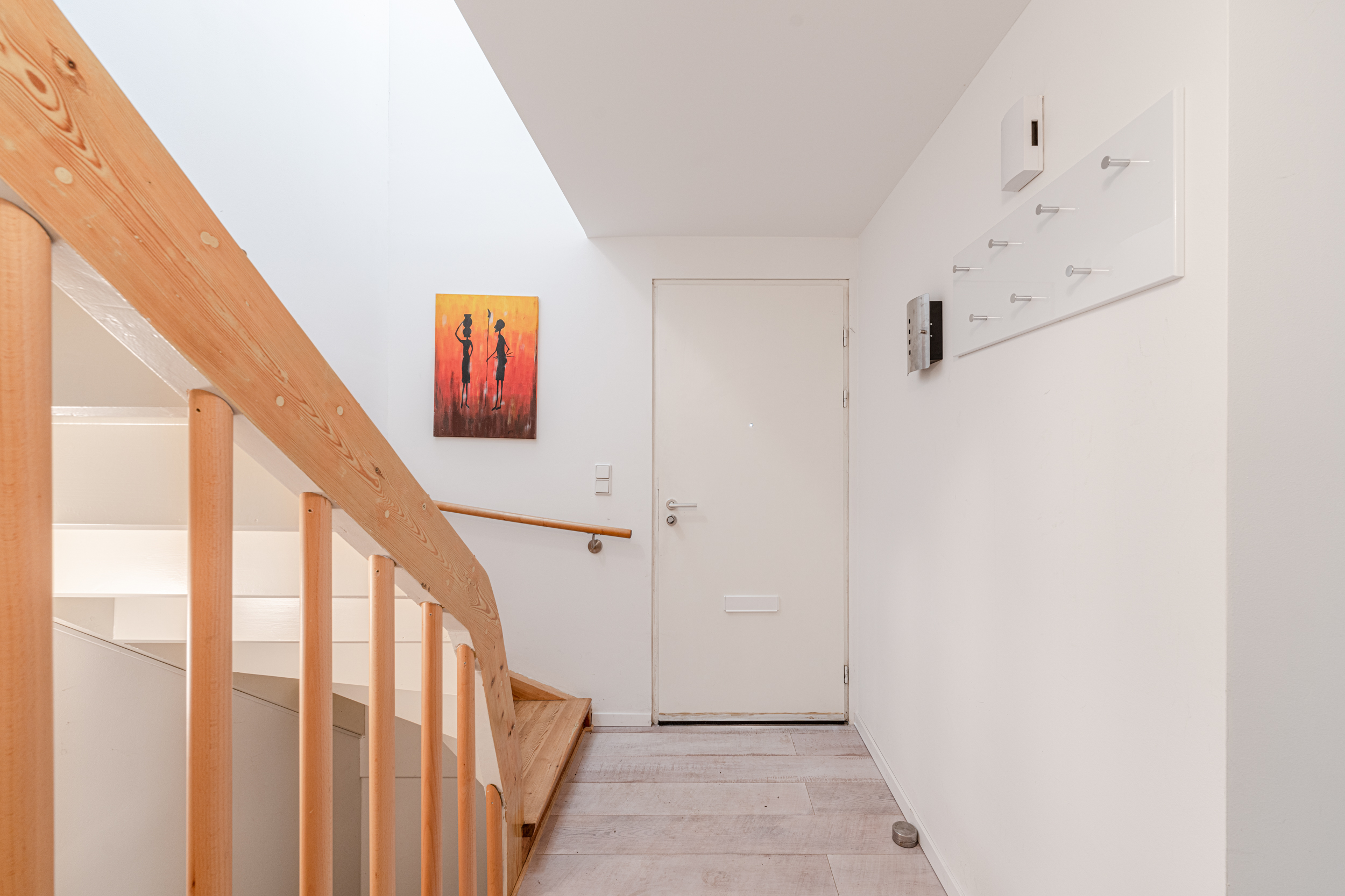 Heller Flur mit Holztreppe, weißer Tür, Wandhaken und farbigem Bild an der Wand.