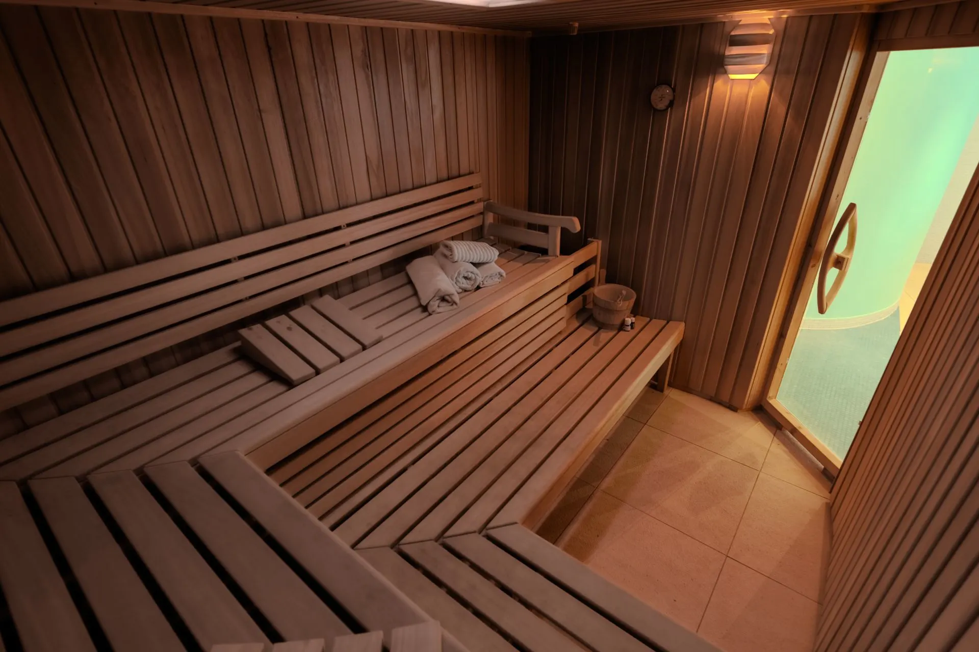Holzsauna mit Bänken, Handtüchern und Kissen, offener Tür und einem Eimer auf einer Bank.