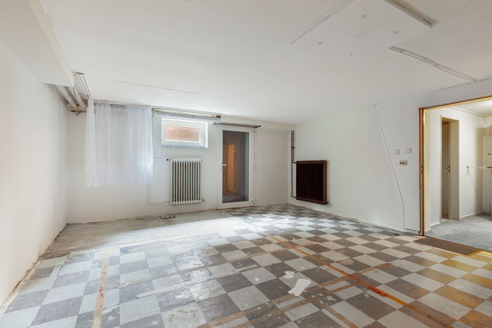 Leerer Kellerraum mit kariertem Boden, Fenster, Glastür und Heizkörper.