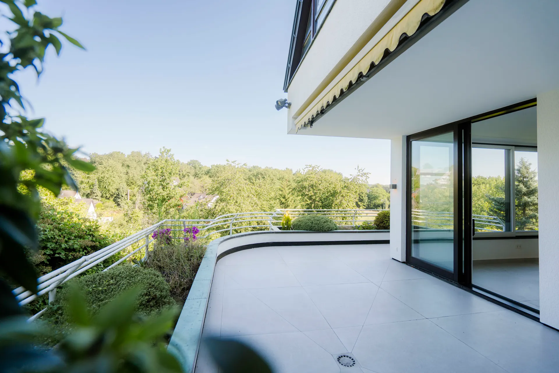 Große Terrasse mit Fliesenboden, Geländer und Blick auf grüne Bäume, angrenzend an ein helles Wohnzimmer.