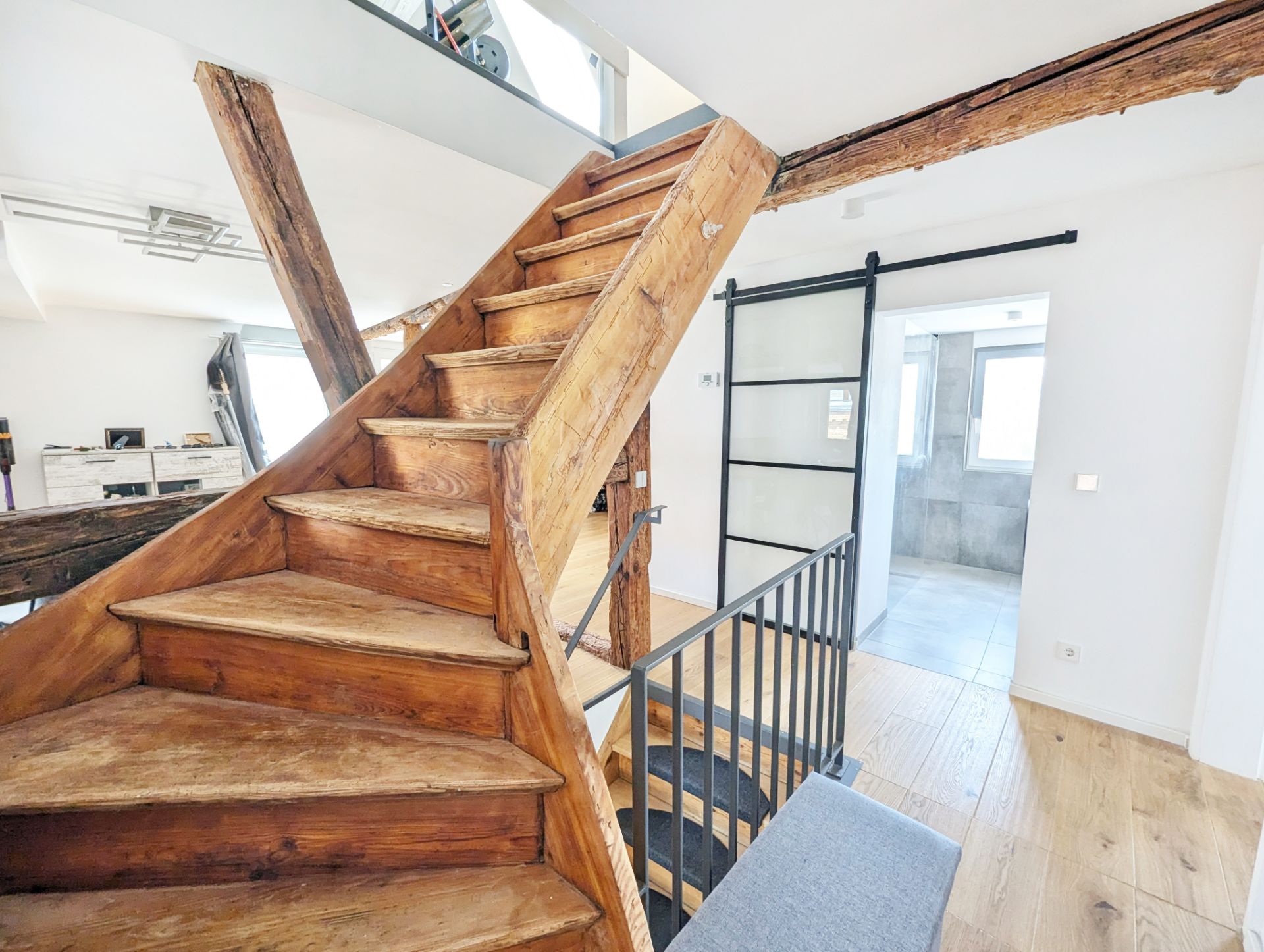 Holztreppe mit sichtbaren Balken in einem hellen Wohnraum, Durchgang mit Schiebetür und Fenster.