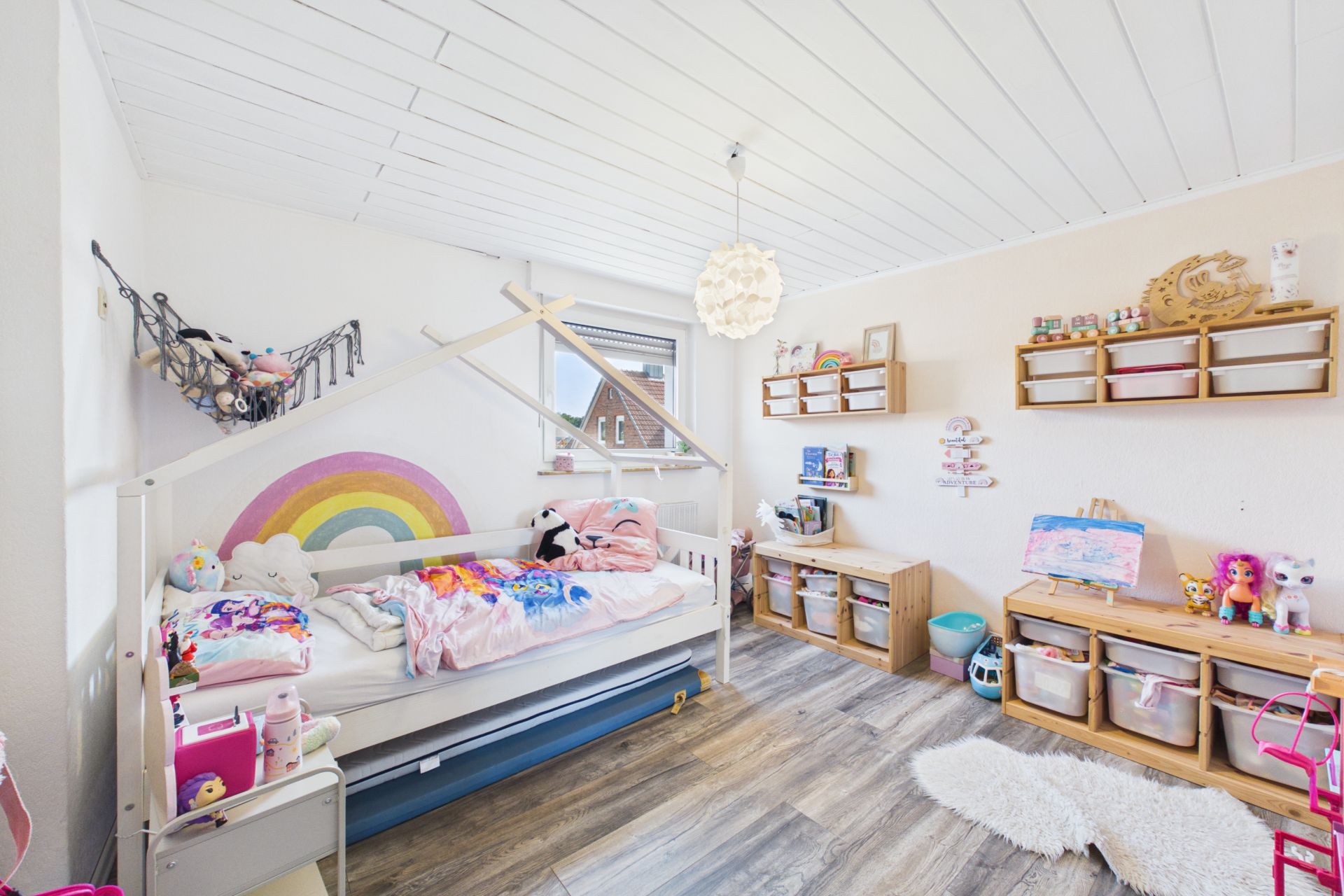 Helles Kinderzimmer mit Holzfußboden, Bett und Regalen voller Spielsachen und Aufbewahrungsboxen.