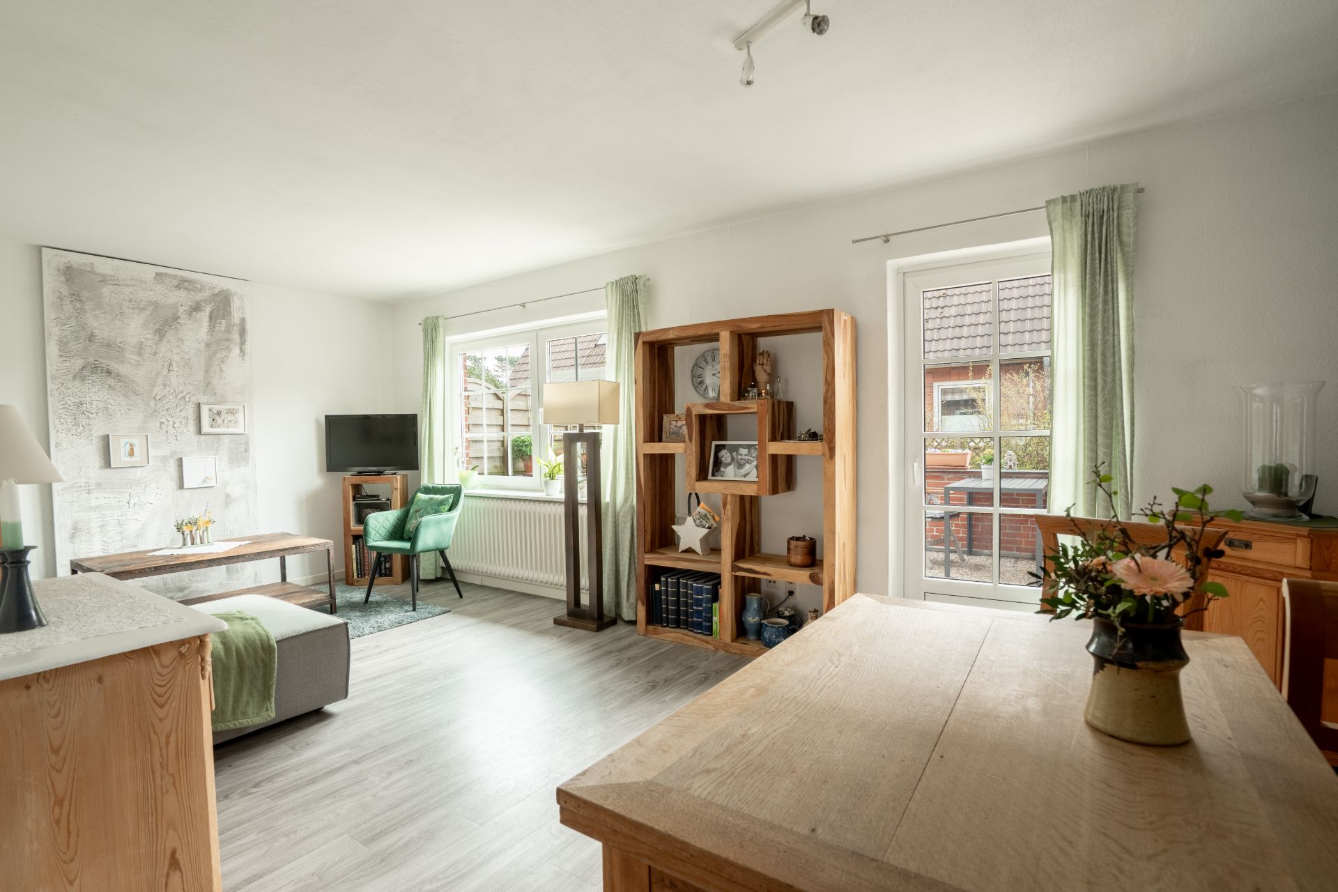 Helles Wohnzimmer mit Holzmöbeln, Sofa, Sessel, Regal, Fenster und Blumen auf dem Tisch.