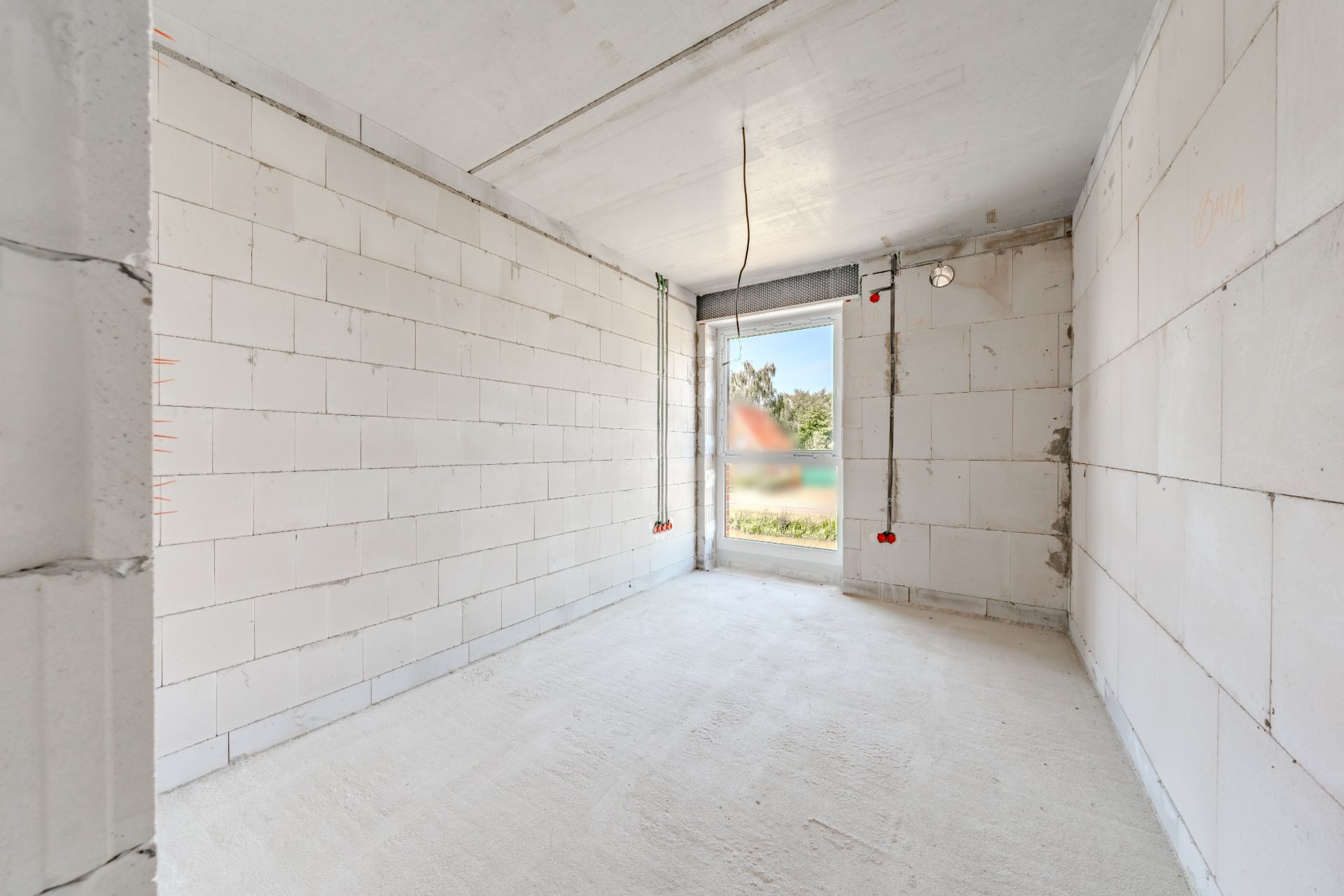 Leerer Raum im Rohbau mit weißen Wänden und großem Fenster am Ende.