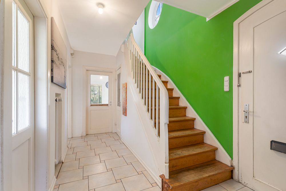 Heller Flur mit Treppe aus Holz und grüner Wand, Türen und gefliestem Boden.