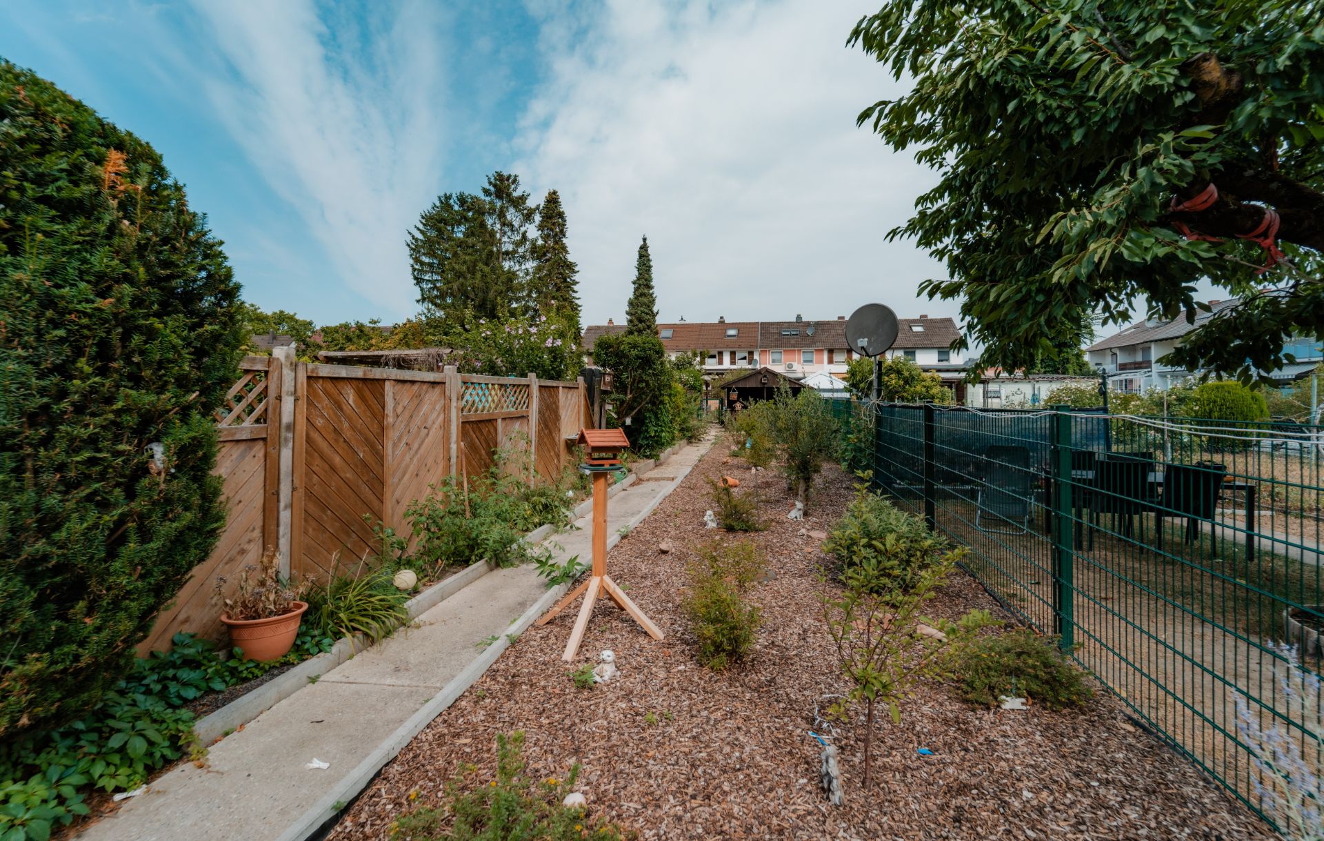 Langer Garten mit Kiesweg, Holzzaun, kleinen Büschen und Vogelhaus, angrenzend an Reihenhäuser.