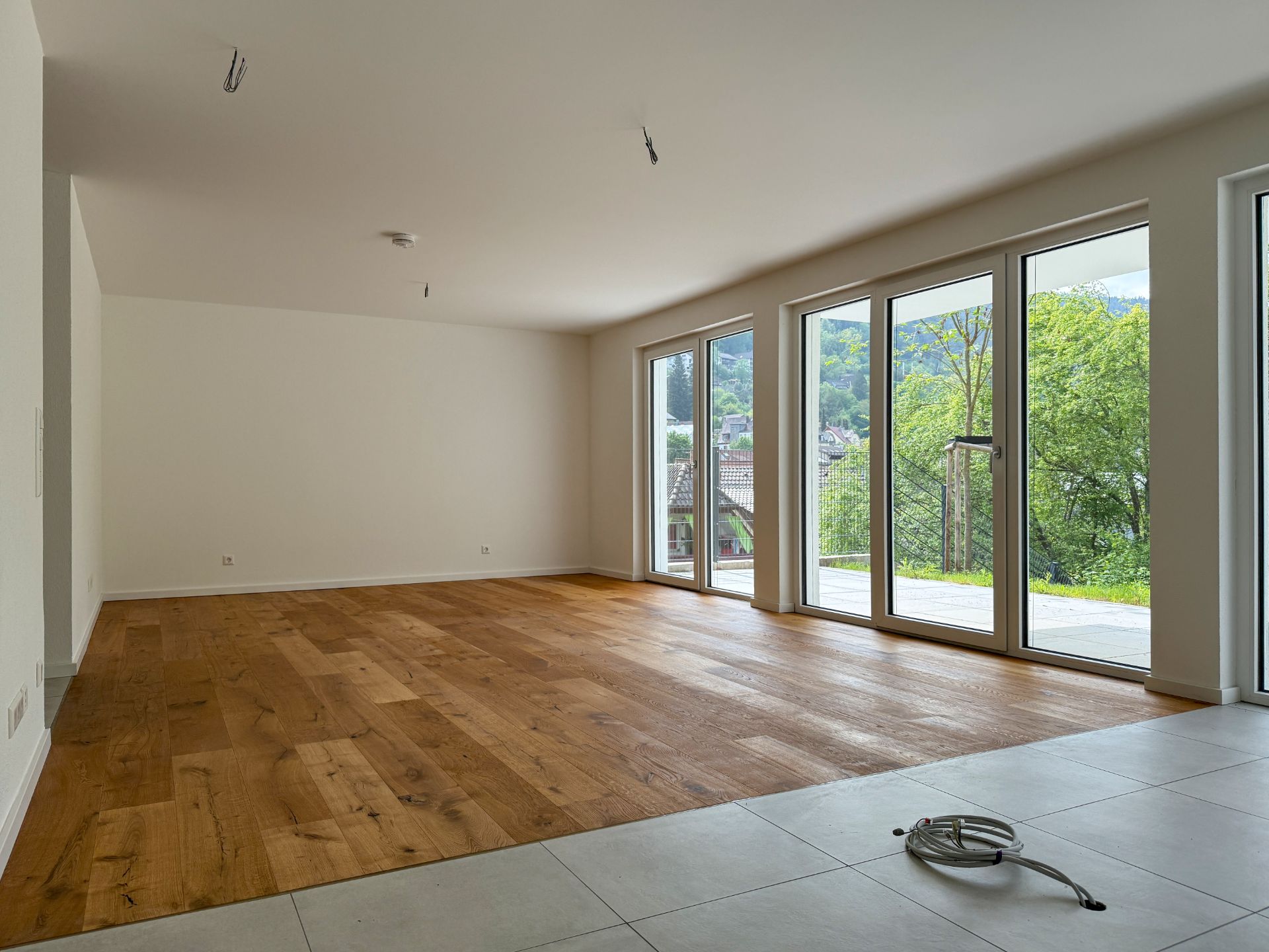 Leerer Wohnraum mit Holzboden und großen Fenstern, Blick auf Terrasse und grüne Landschaft.