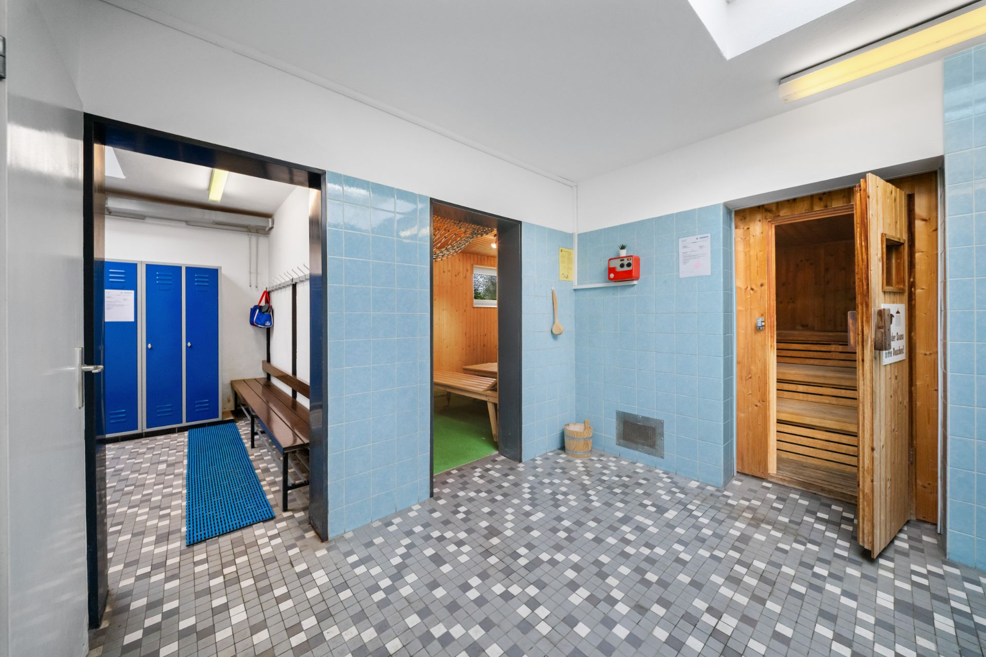 Umkleideraum mit blauen Spinden, Holzbänken und geöffneter Tür zu einer Sauna.