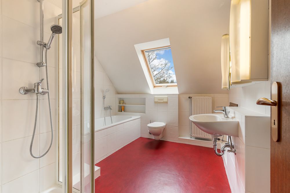 Helles Badezimmer mit rotem Boden, Dusche, Badewanne, WC, Waschbecken und Dachfenster.