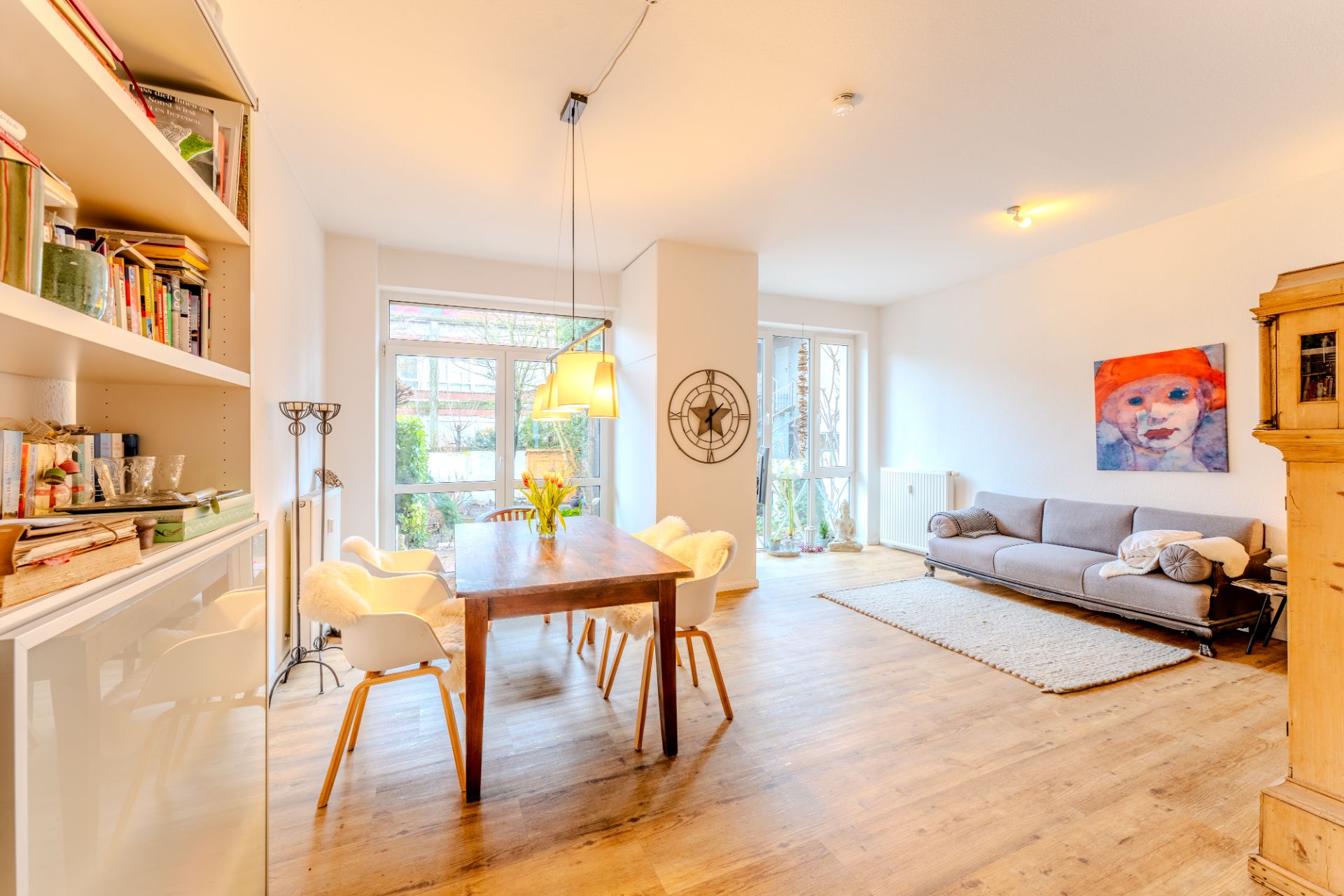 Helles Wohnzimmer mit Holzboden, Esstisch, Regal, Sofa, Teppich und großen Fenstern.