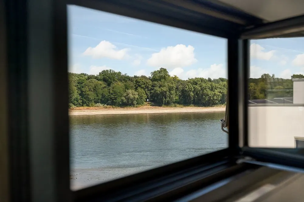 Blick durch ein Fenster auf einen Fluss und eine bewaldete Uferlandschaft.