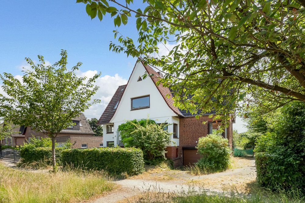 Zweistöckiges Haus mit Satteldach, umgeben von Bäumen und Sträuchern an einer Einfahrt.