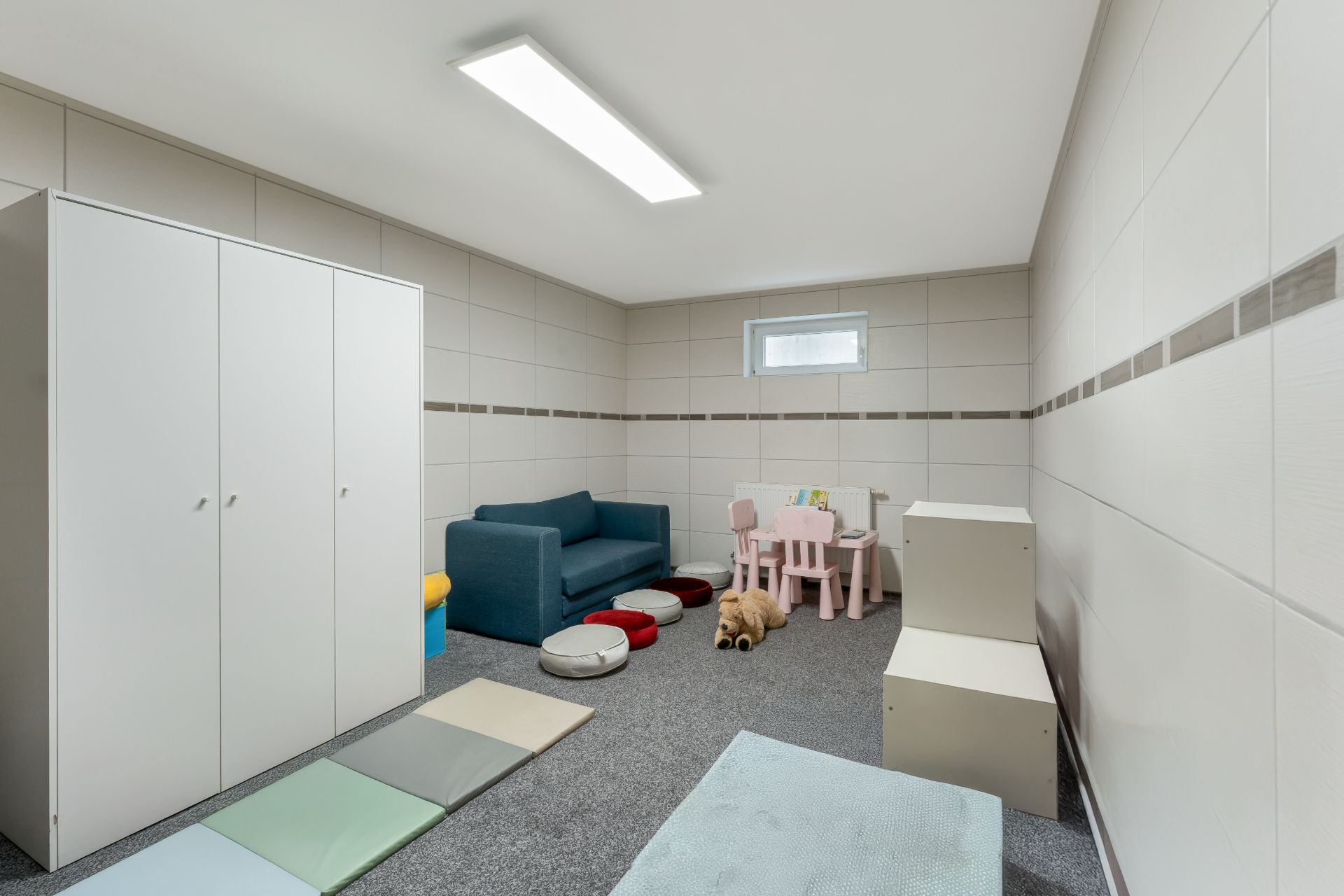 Heller Raum mit Sofa, Spielmatten, Kindertisch, Stühlen und Schrank, Spielzeug auf dem Boden.