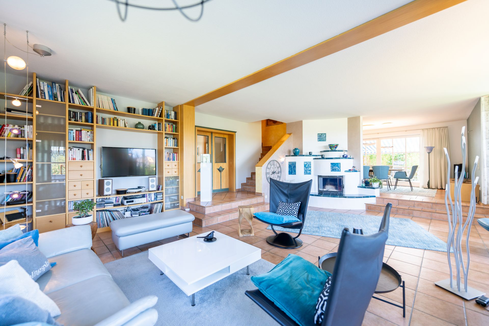 Helles Wohnzimmer mit Sofas, Sessel, TV-Regalwand, Kamin und offener Treppe.