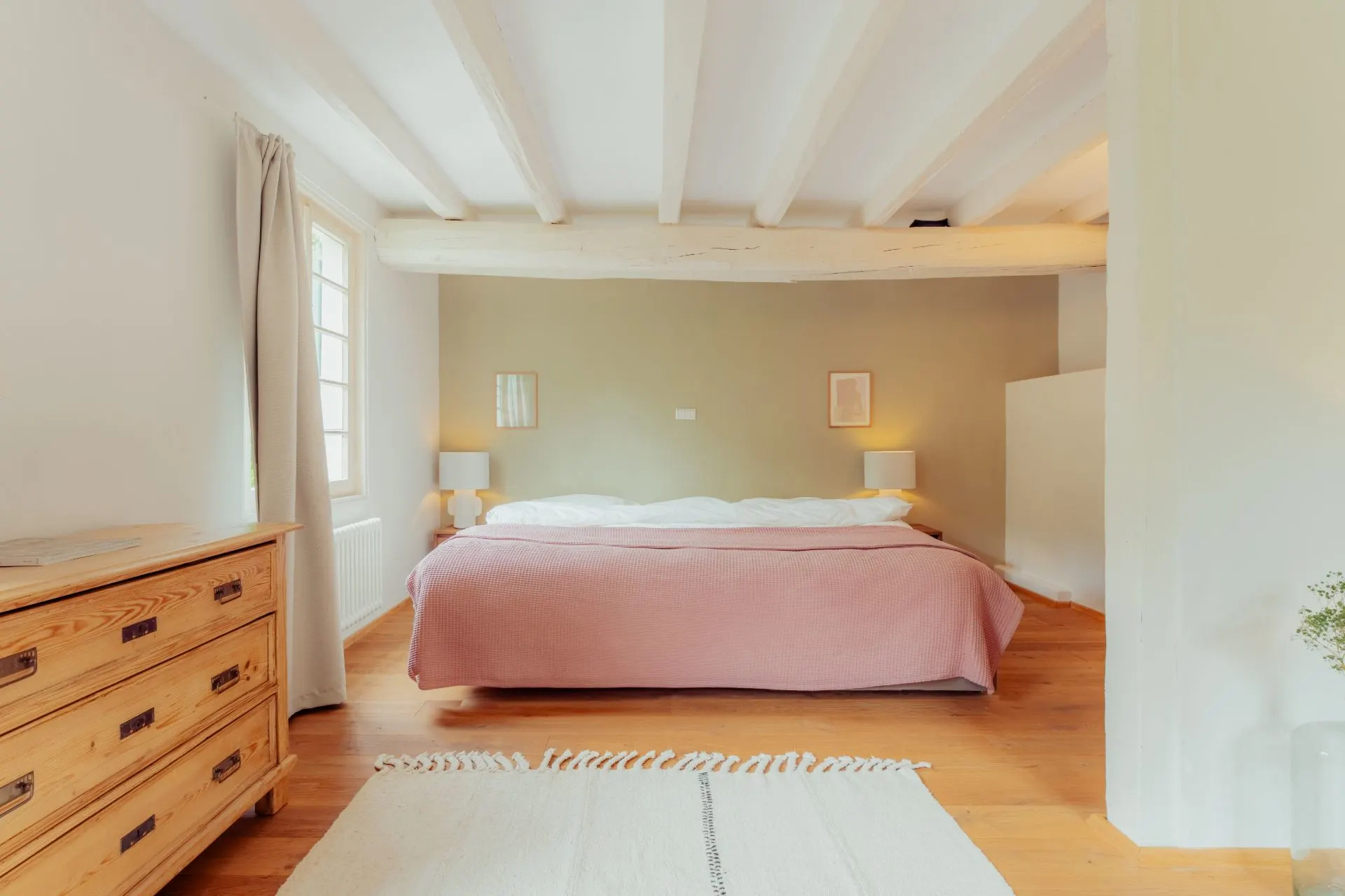 Helles Schlafzimmer mit Doppelbett, rosa Decke, Holzboden und Holzbalken an der Decke.