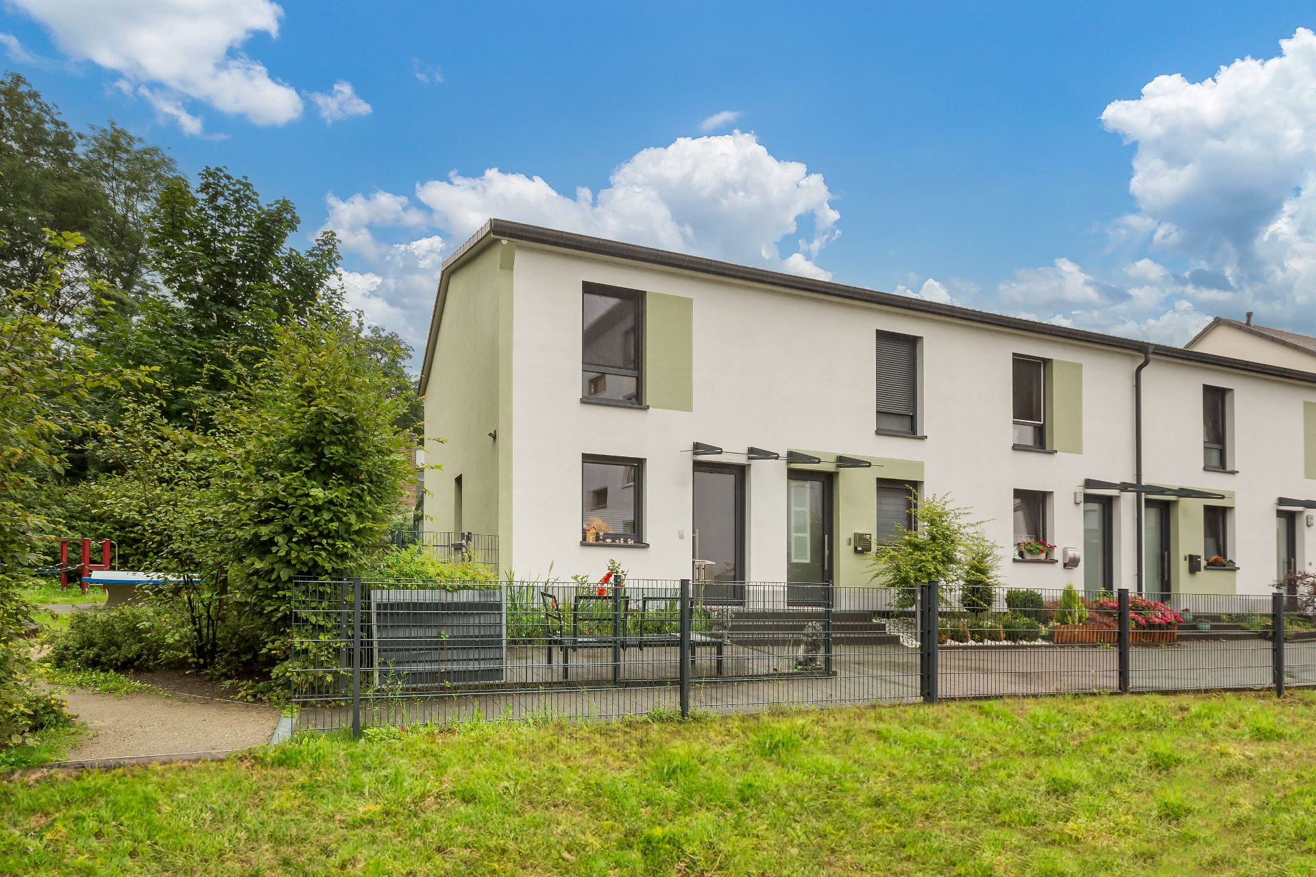 Reihenhaus mit Garten, Zaun und Terrasse, im Vordergrund Rasen, im Hintergrund Bäume und blauer Himmel.