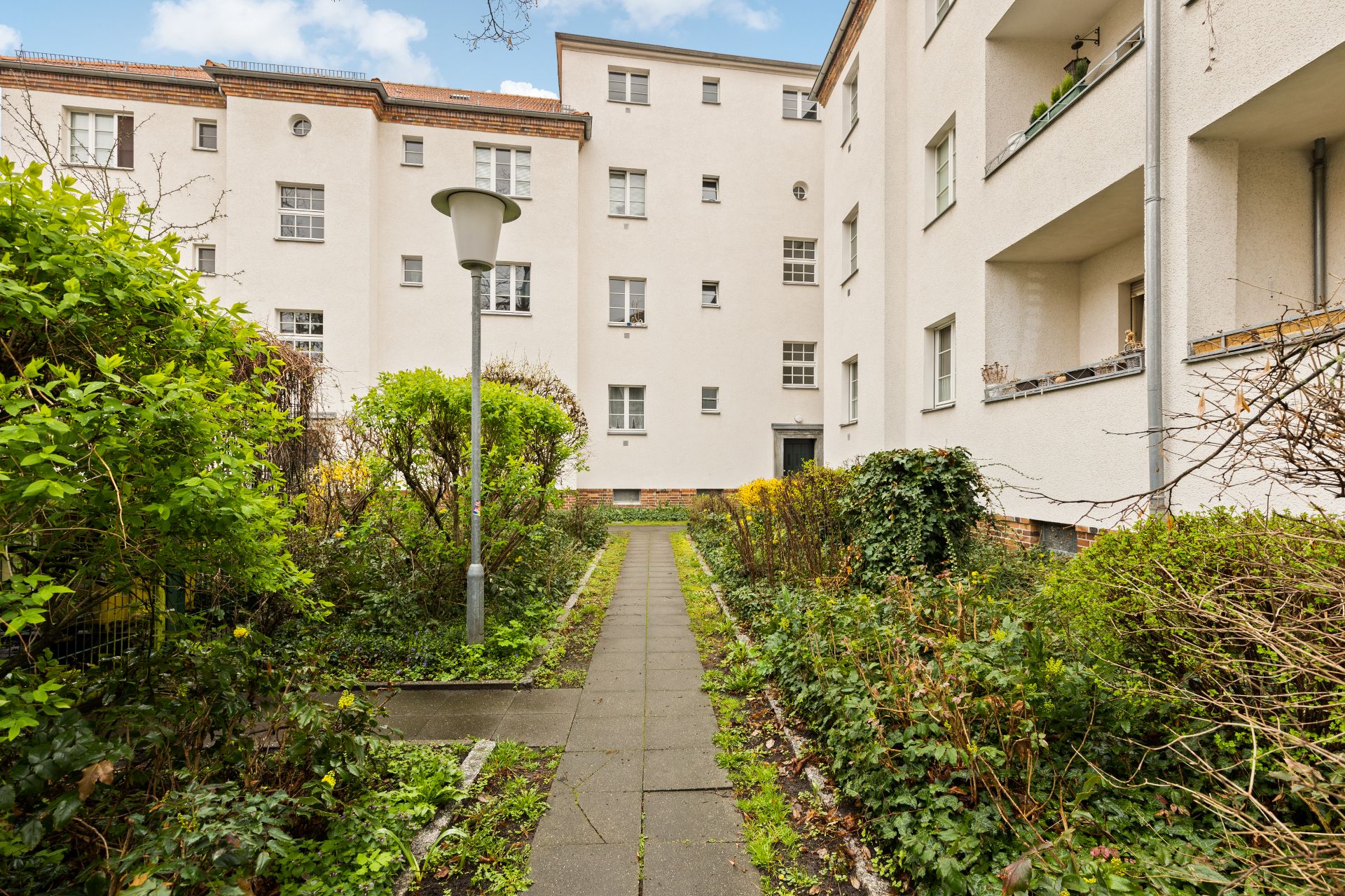 Gepflegter Innenhof mit Gehweg, Laterne, Sträuchern und weißen Mehrfamilienhäusern.