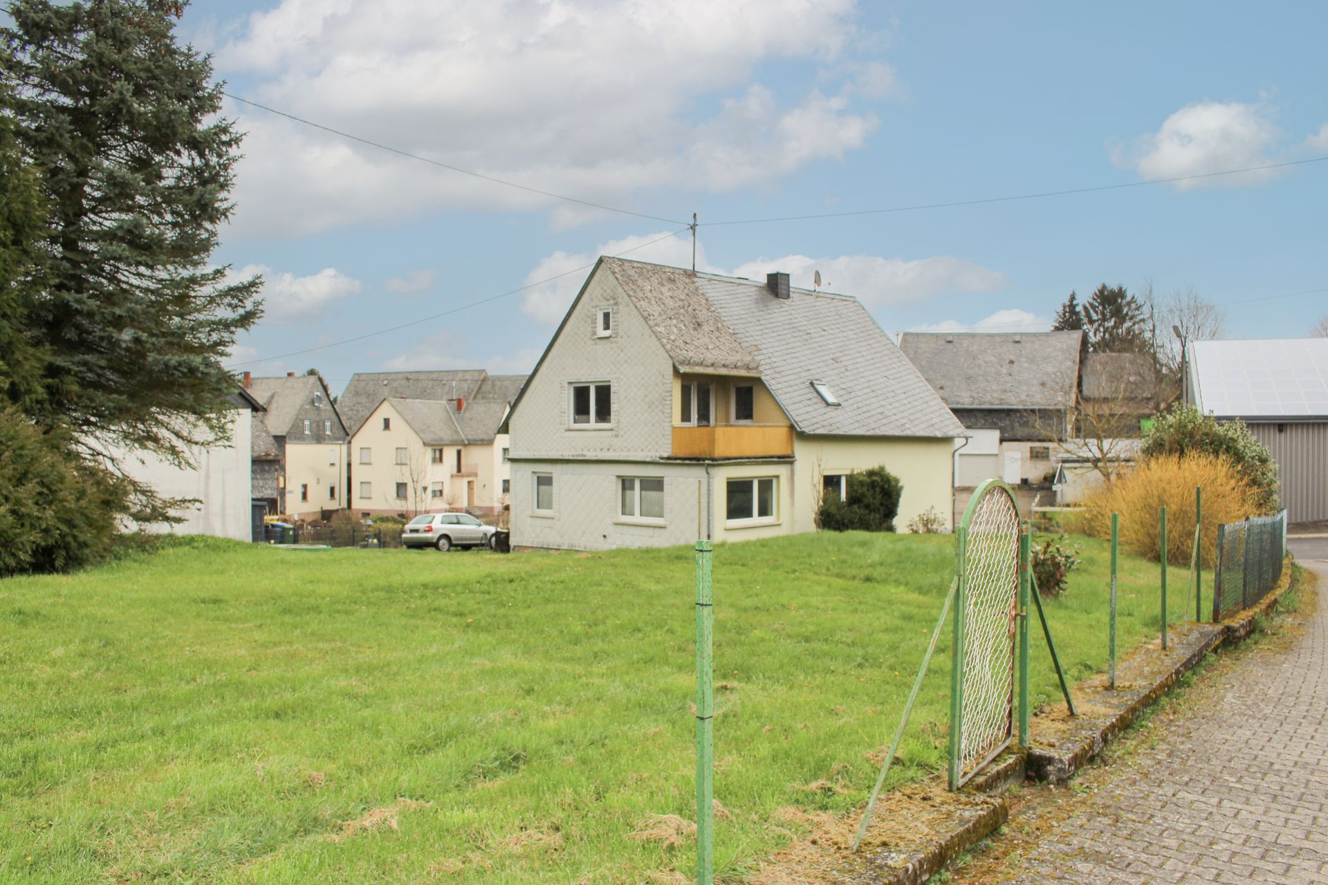 Wohnhaus mit spitzem Dach, großem Garten und grünem Zaun bei Tageslicht.