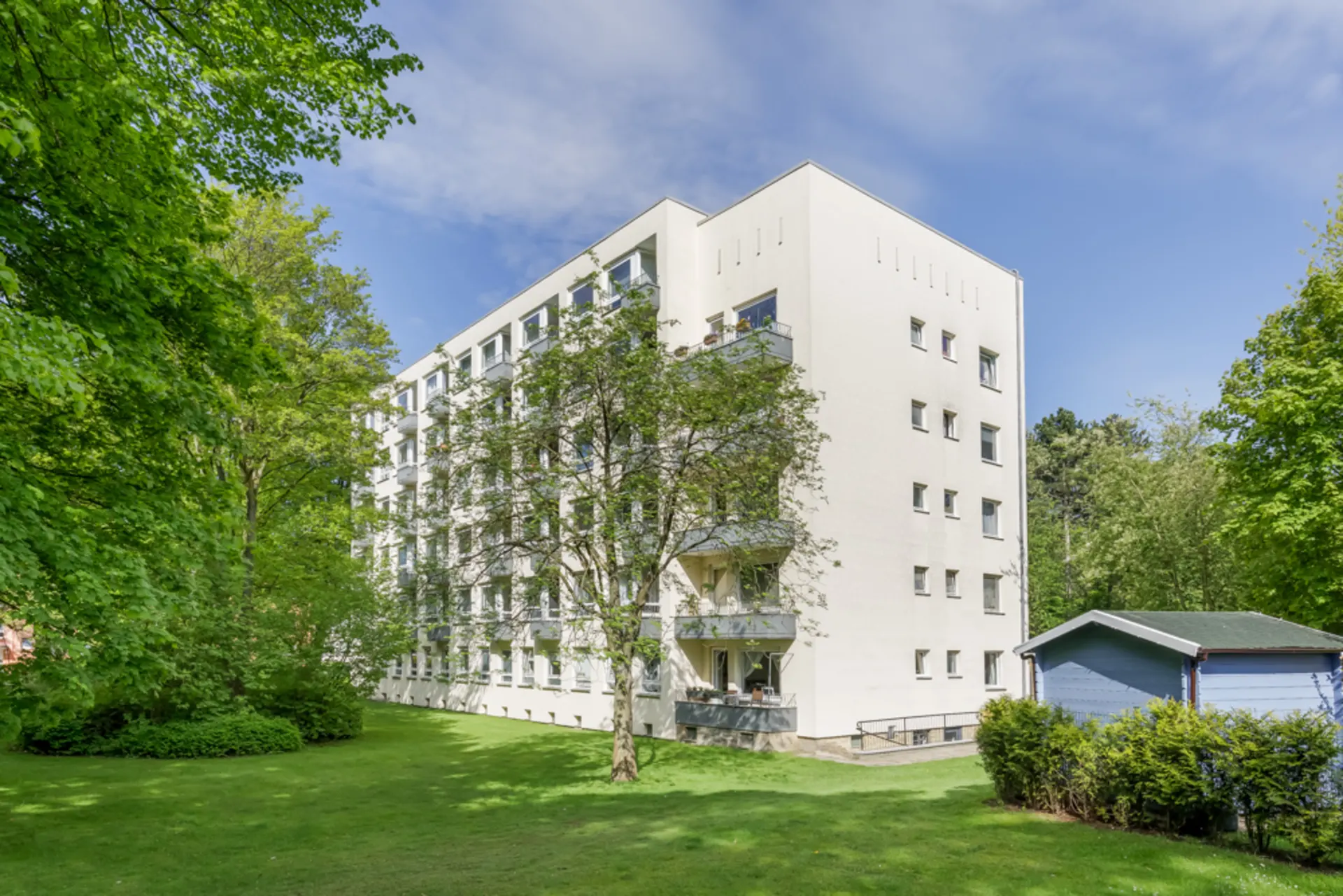 Mehrgeschossiges Wohnhaus mit Balkonen, umgeben von Bäumen und einer großen Wiese.