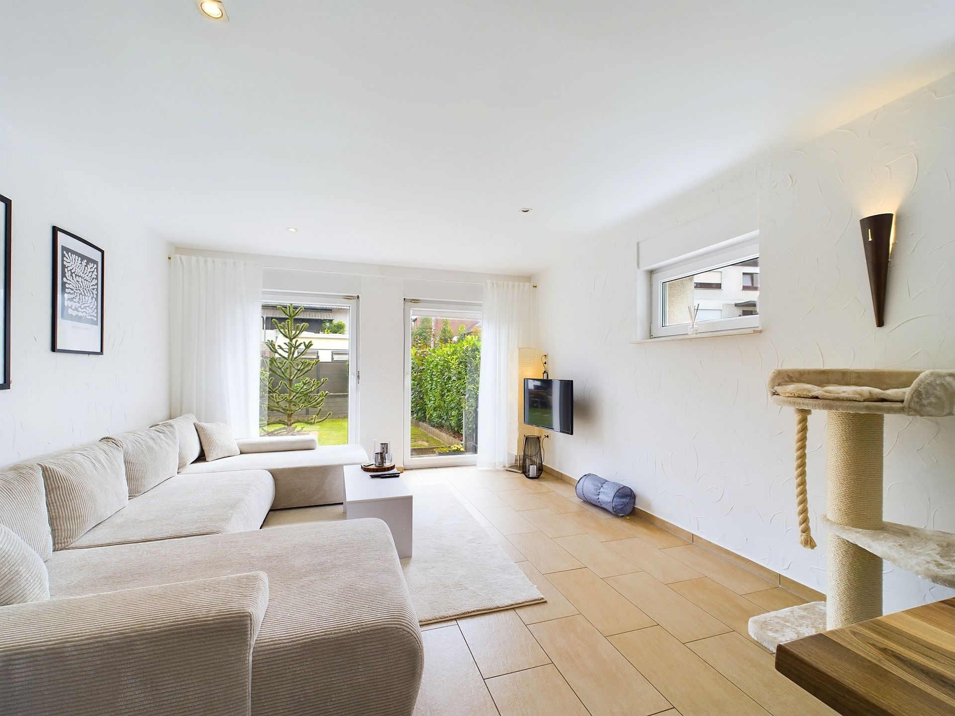 Helles Wohnzimmer mit beigem Sofa, Glastür zum Garten und Kratzbaum an der Wand.