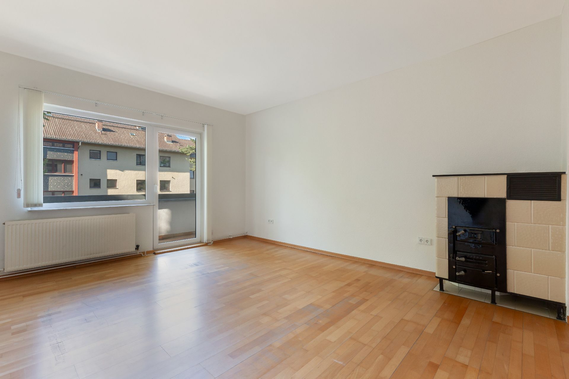 Leeres Zimmer mit Holzfußboden, großem Fenster, Heizkörper und Kaminofen an der Wand.