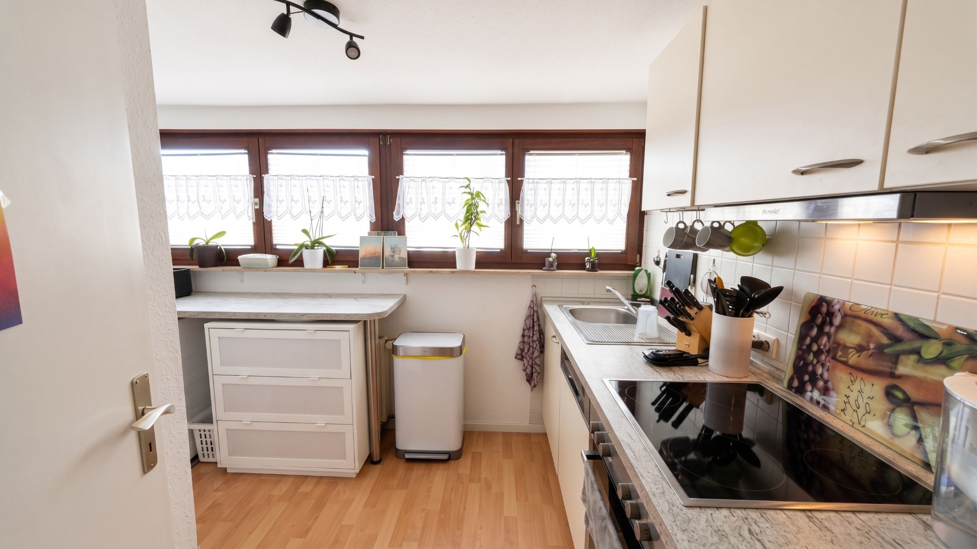 Helle Küche mit Holzfußboden, weißen Schränken und großen Fenstern mit Pflanzen auf der Fensterbank.