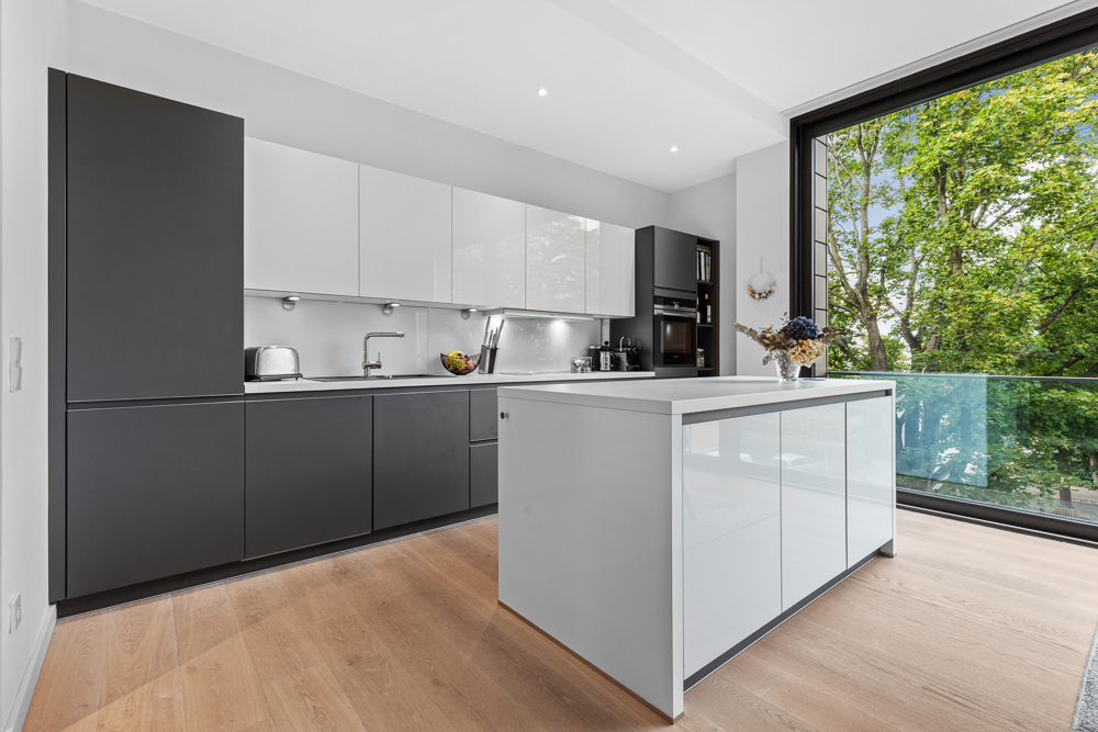 Moderne Küche mit grauen und weißen Schränken, Kücheninsel und großem Fenster mit Blick auf Bäume.