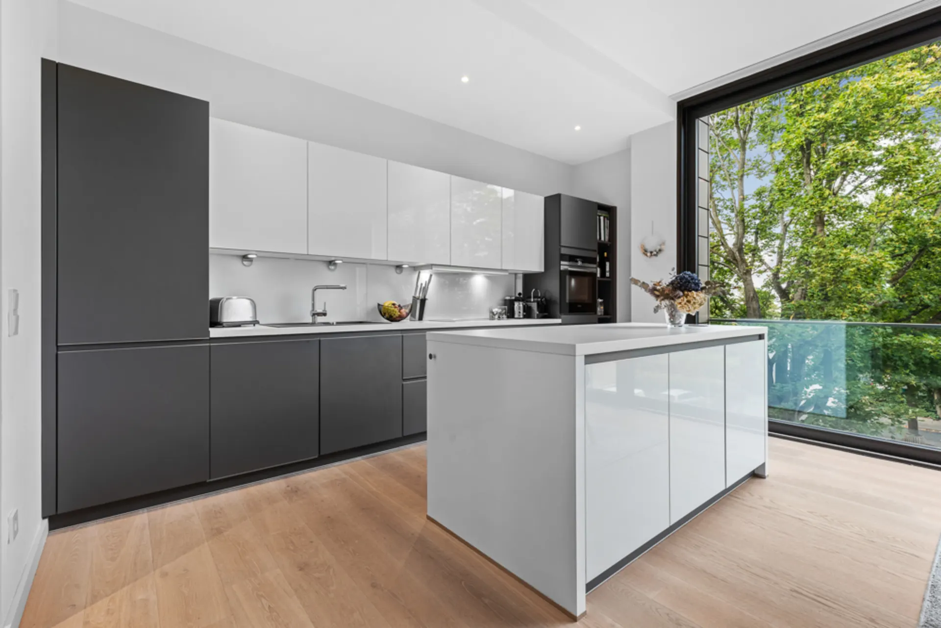 Moderne Küche mit grauen und weißen Schränken, Kücheninsel und großem Fenster mit Blick auf Bäume.
