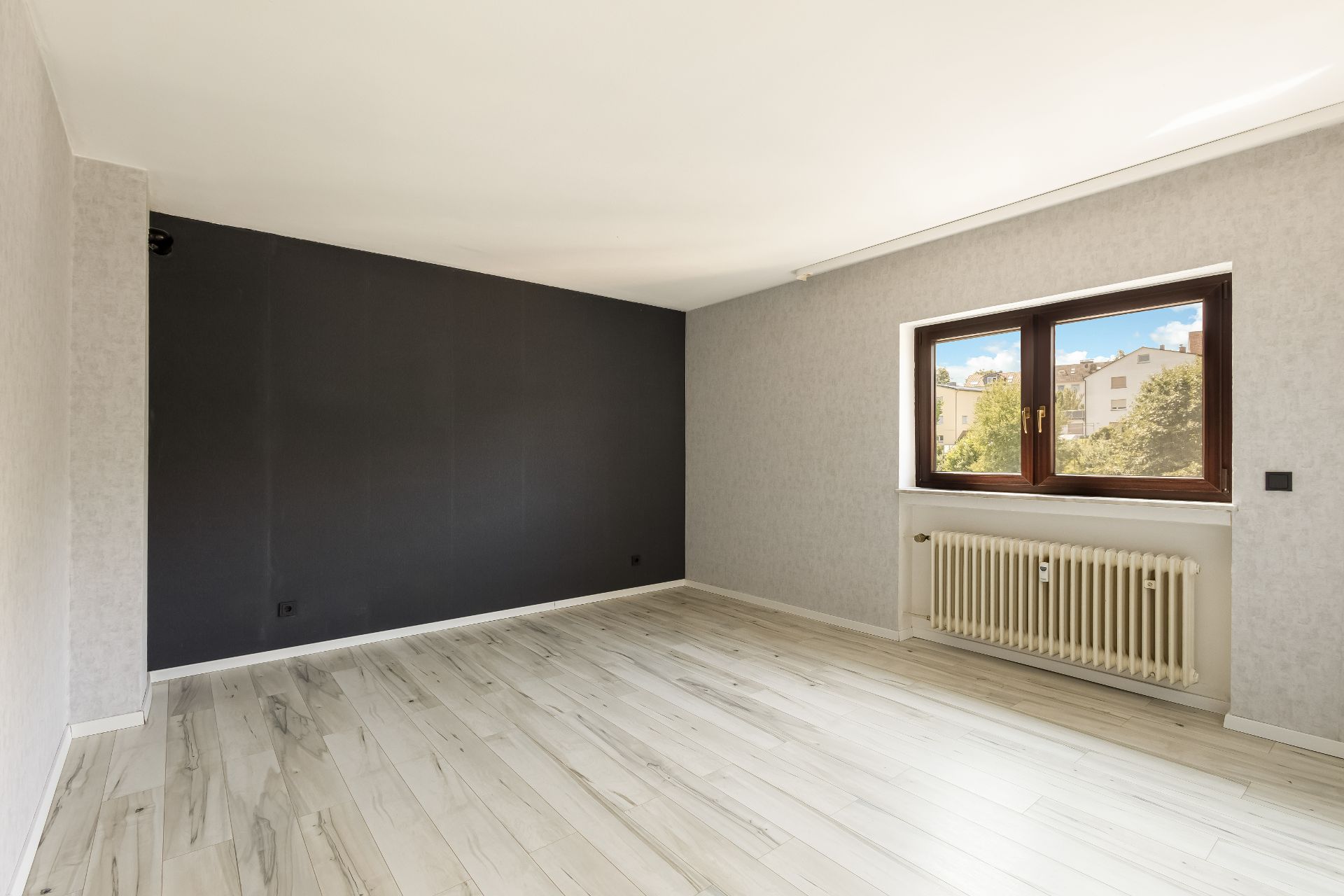 Leerer Raum mit hellem Boden, dunkler Wand und großem Fenster mit Blick ins Freie.