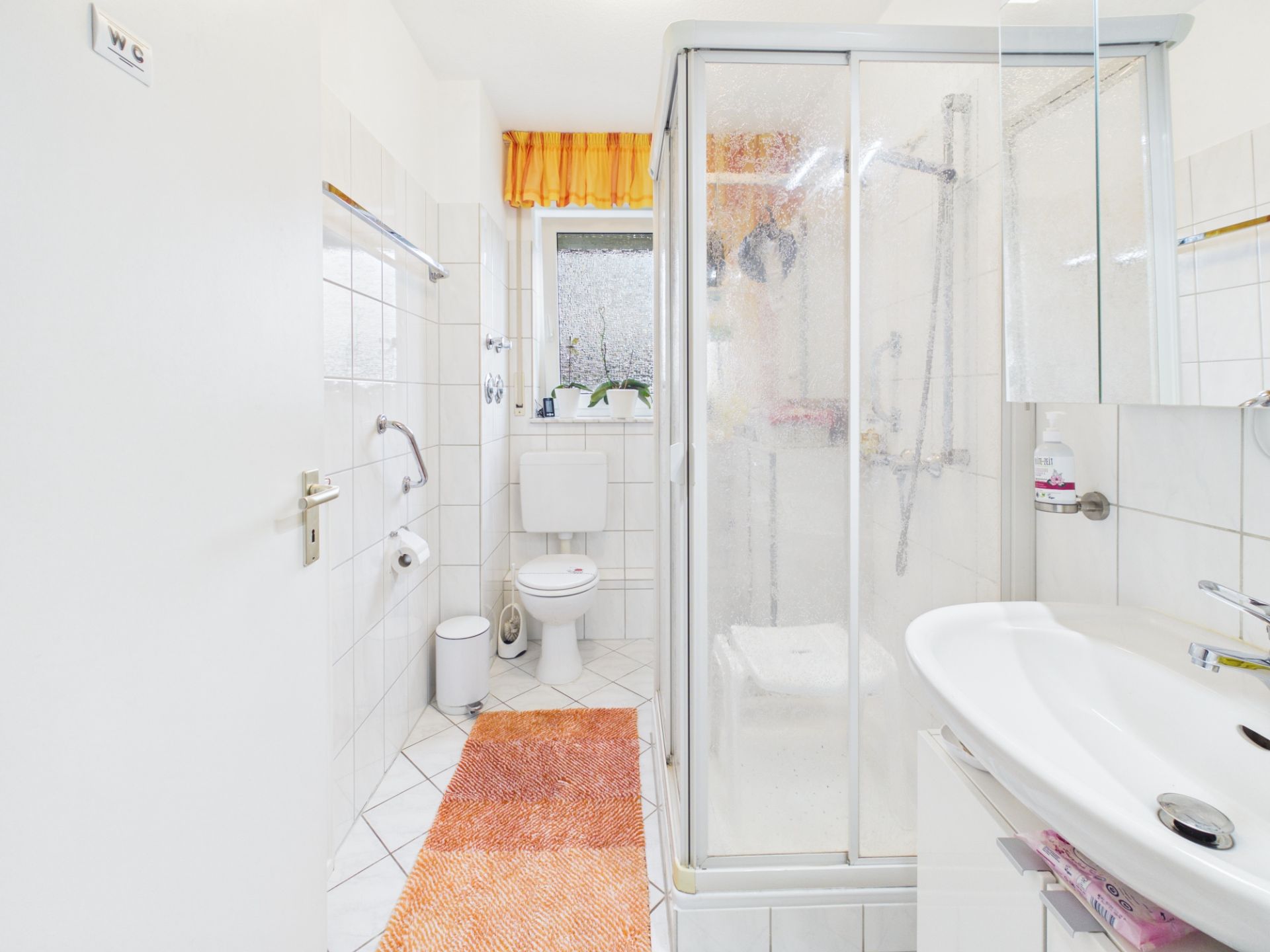 Helles Badezimmer mit Dusche, Toilette, Waschbecken und orangefarbenem Teppich.