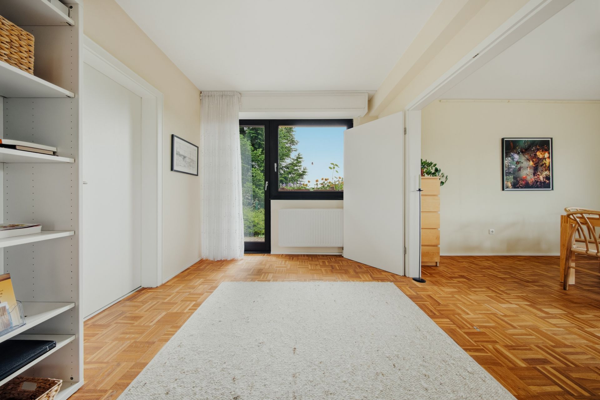 Helles Zimmer mit Parkettboden, Teppich, Regal, großem Fenster und Blick ins Grüne.