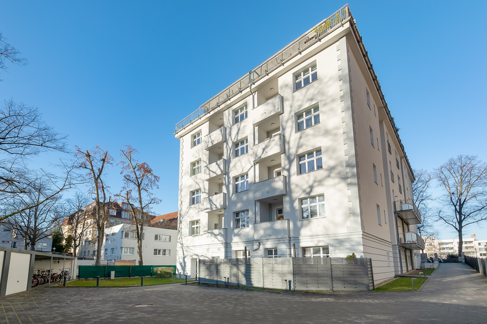 Mehrstöckiges Wohnhaus mit Balkonen und mehreren Fenstern, umgeben von Bäumen und Gehweg.